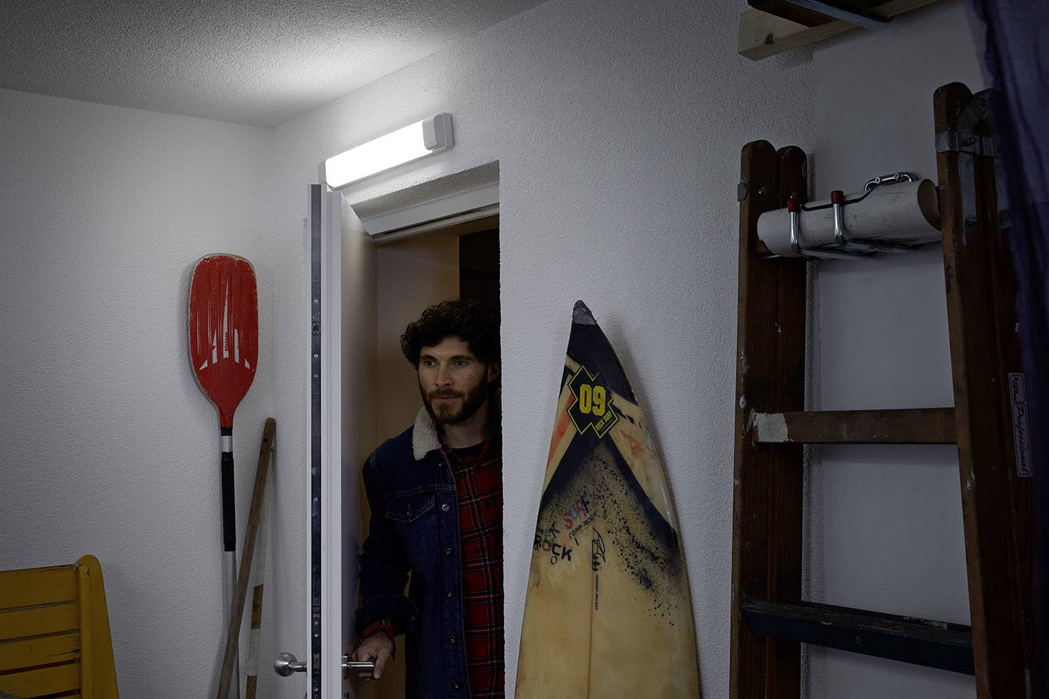 Ein Mann in Jeansjacke betritt einen Abstellraum mit Leiter, Paddel und Surfbrett. Eine Lampe leuchtet den Raum aus.