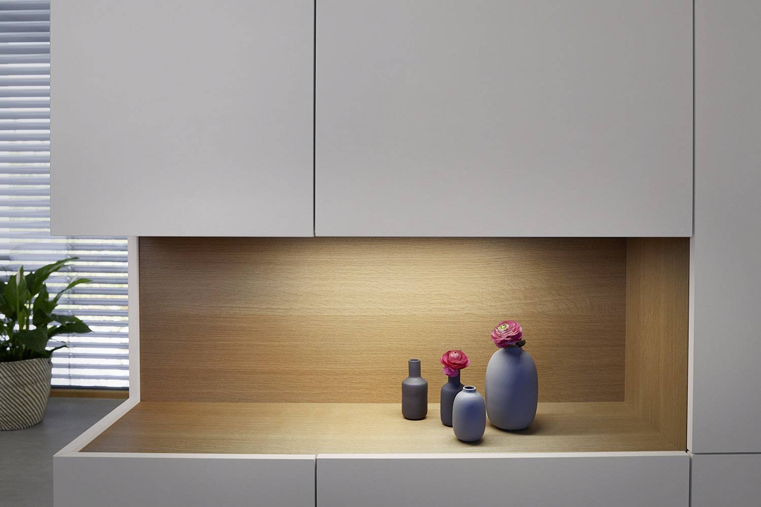 Dekorative Nische in einem Schrank mit drei grauen Vasen und rosafarbenen Blumen, sanft beleuchtet auf einem Holzregal.