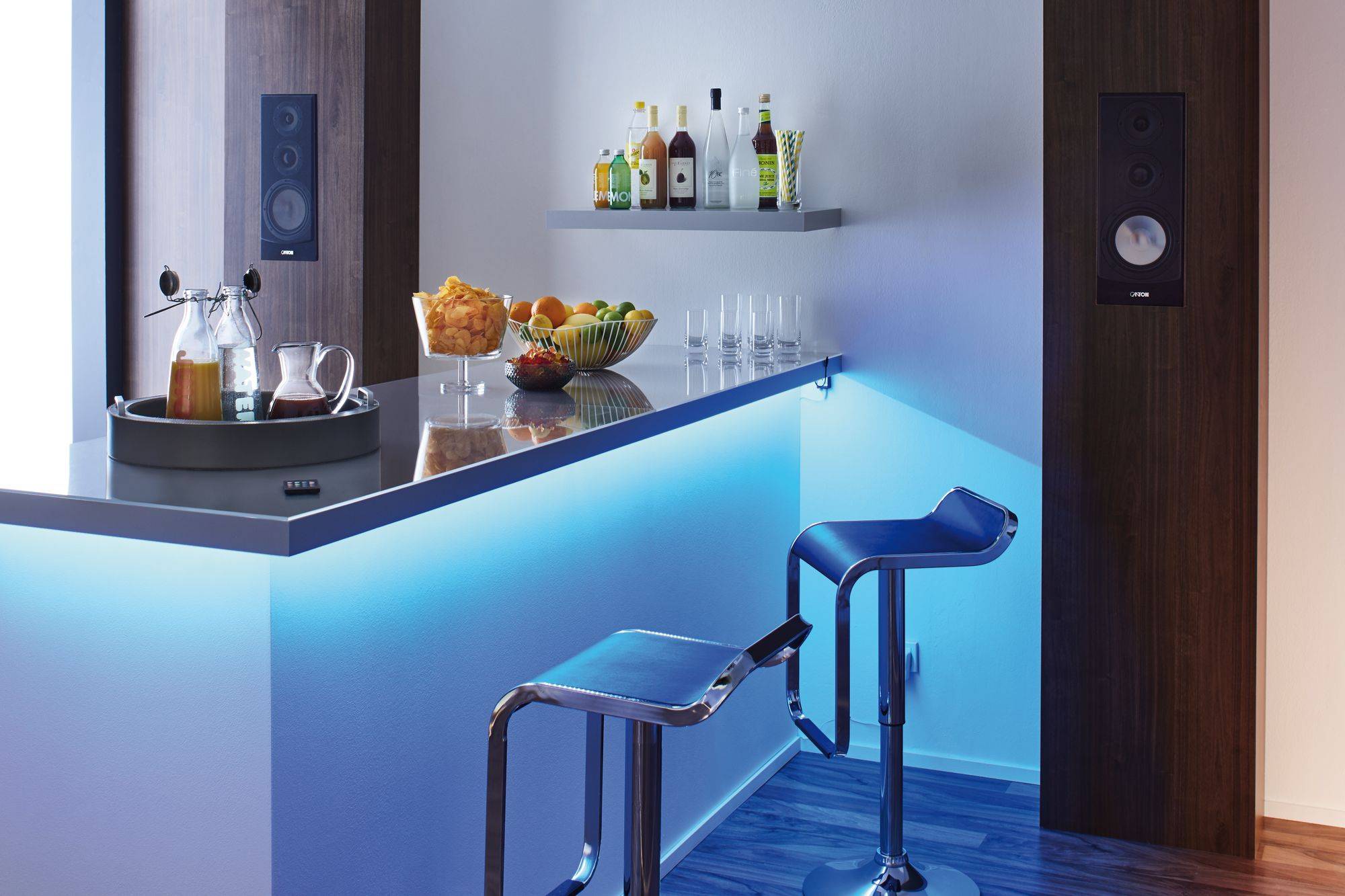 Moderne Hausbar mit blauer LED-Beleuchtung. Zwei Barhocker, Flaschen und Gläser im Hintergrund auf einem Regal.