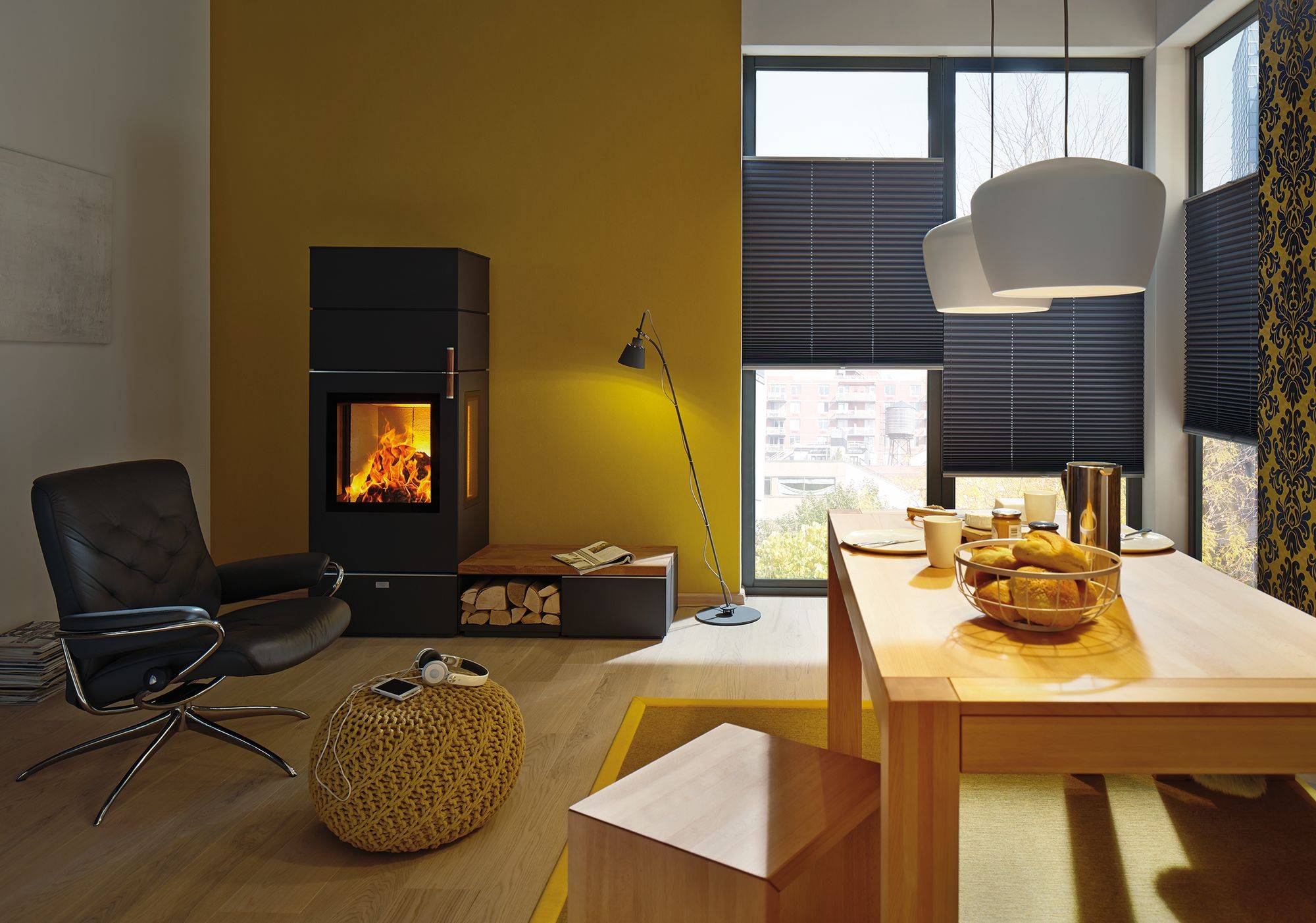 Modernes Wohnzimmer mit gelber Wand und schwarzen Möbeln. Kamin an, stimmungsvolle Beleuchtung. Holztisch mit Frühstücksplatte. Gemütliche Atmosphäre.