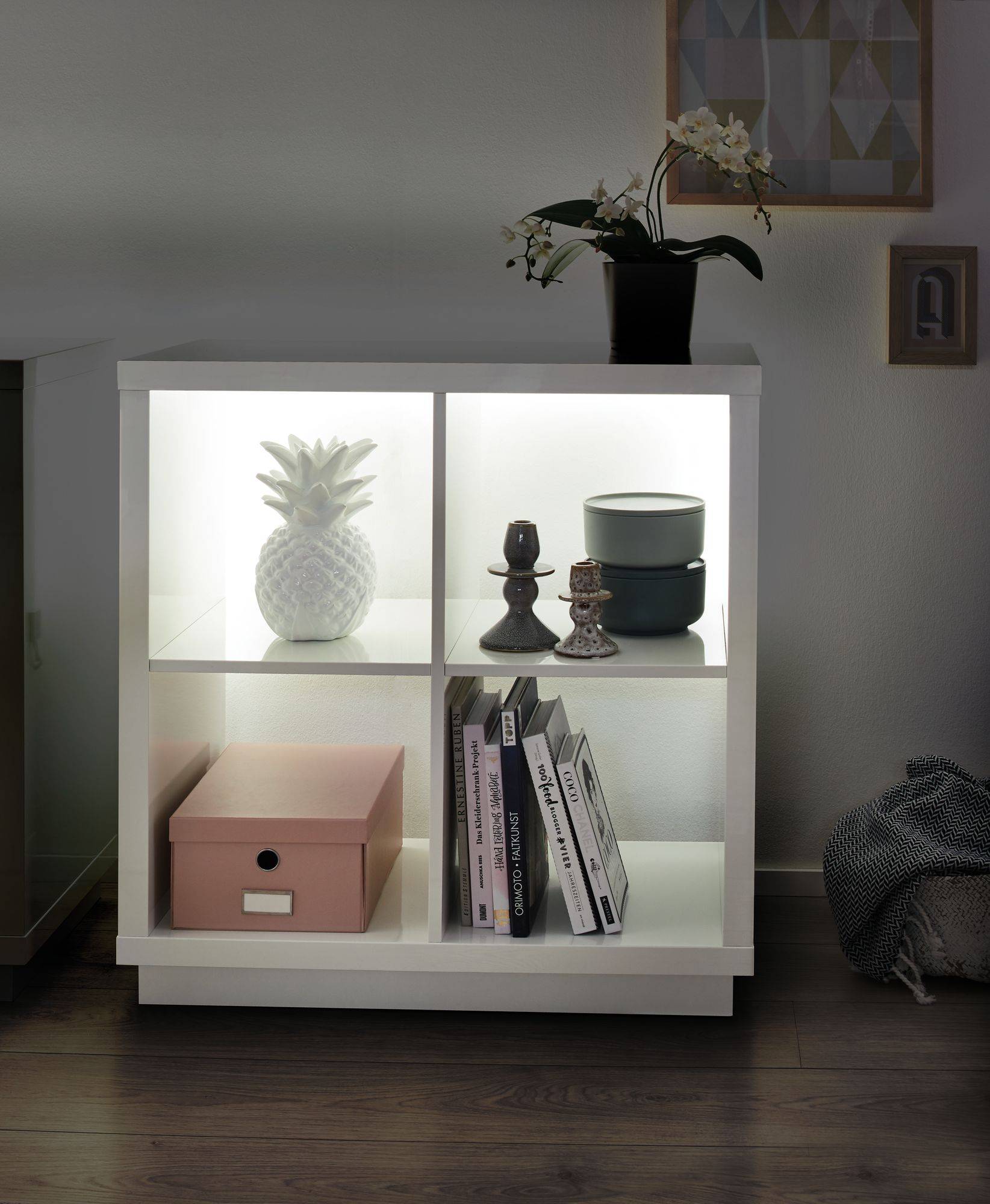 Weißes Regal mit Beleuchtung, darin eine weiße Ananas, Buchstützen, Bücher und eine pinke Box. Oben eine Orchidee.