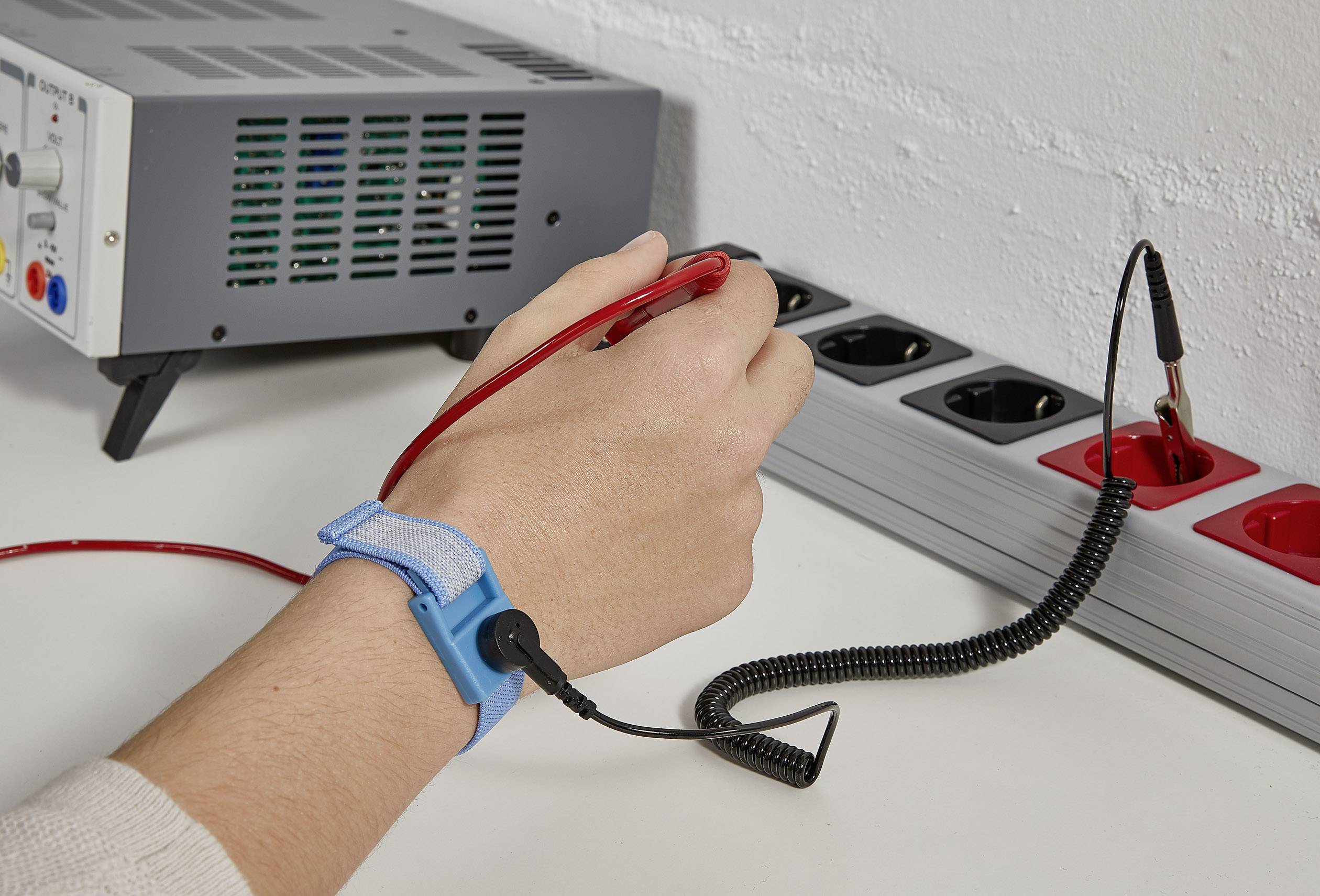 Eine Hand trägt ein antistatisches Armband und hält eine rote Prüfsonde, die in eine Steckdose eingeführt wird. Ein Strommessgerät steht im Hintergrund.