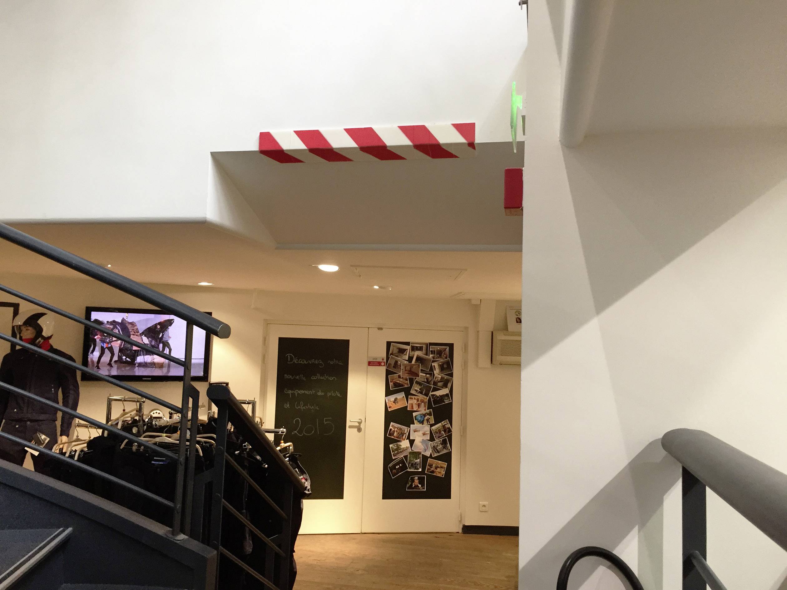 Treppenhaus mit schwarzem Geländer, im Hintergrund eine Tür mit Fotos beklebt und ein Fernseher an der Wand, zeigt Menschen.