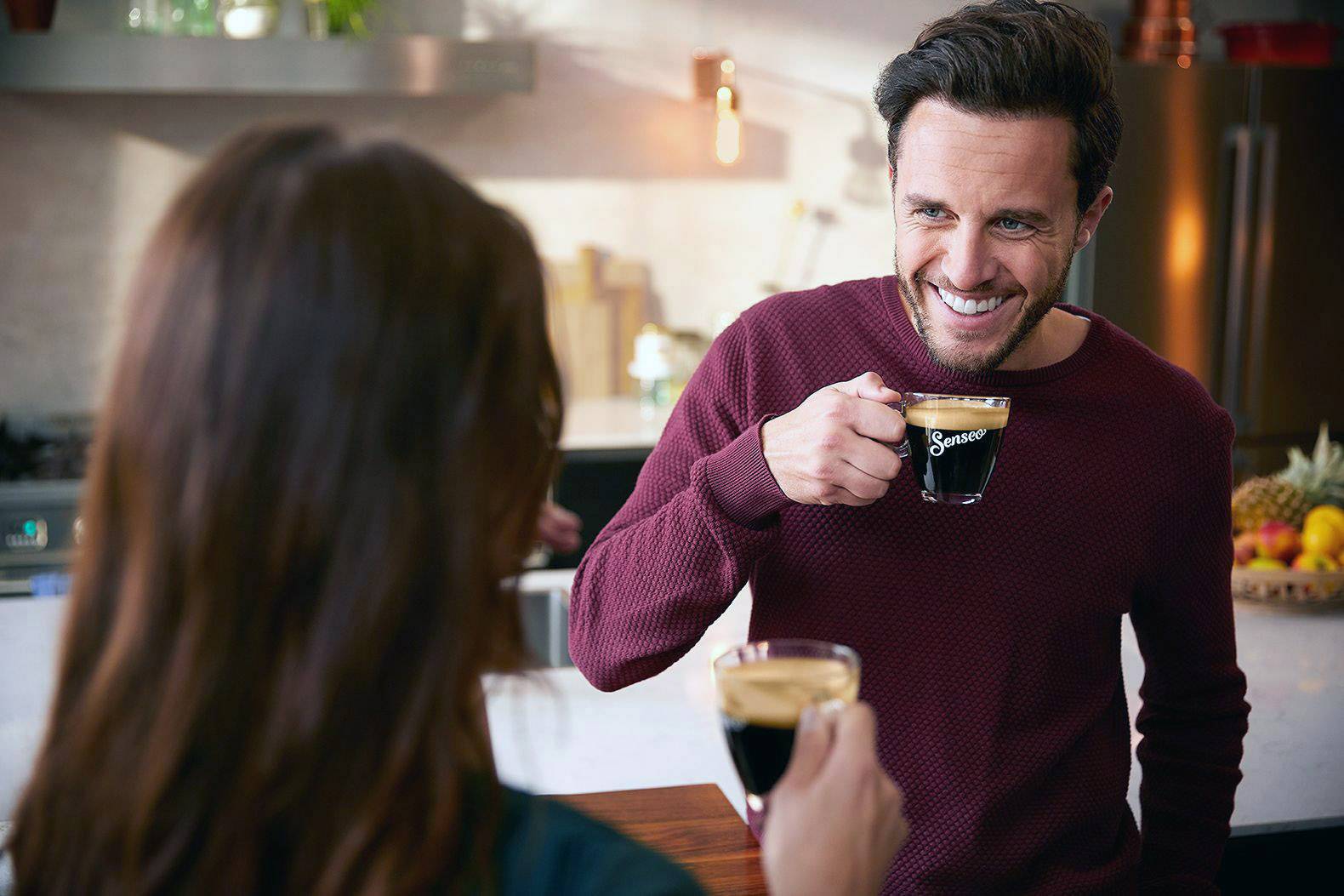 Ein Mann lächelt und hält eine Tasse Kaffee in einer modernen Küche. Gegenüber steht eine Frau, die ebenfalls Kaffee hält.