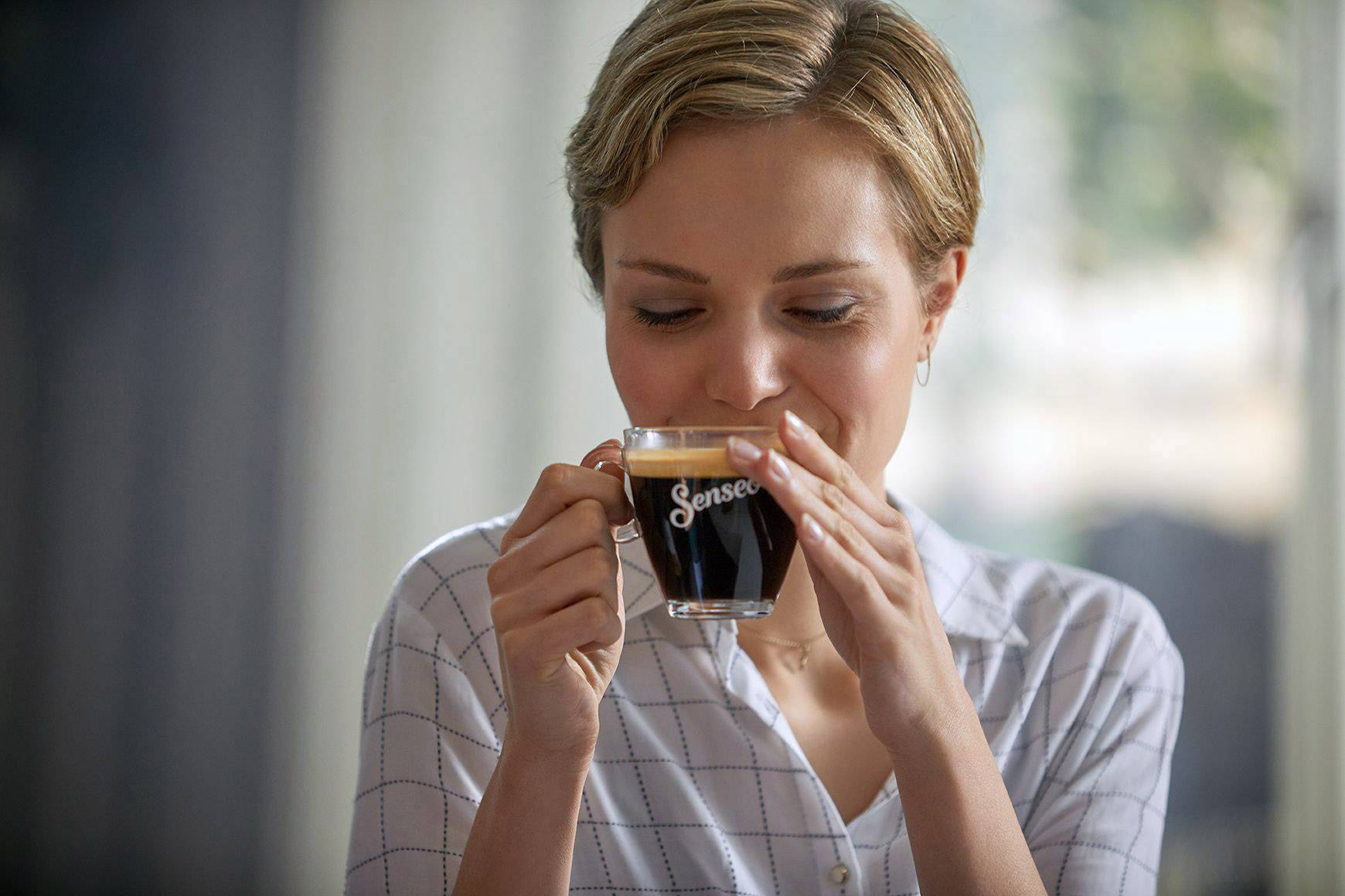 Eine Person trinkt aus einer kleinen Senseo-Tasse Kaffee und lächelt dabei leicht. Im Hintergrund verschwommenes Interieur.