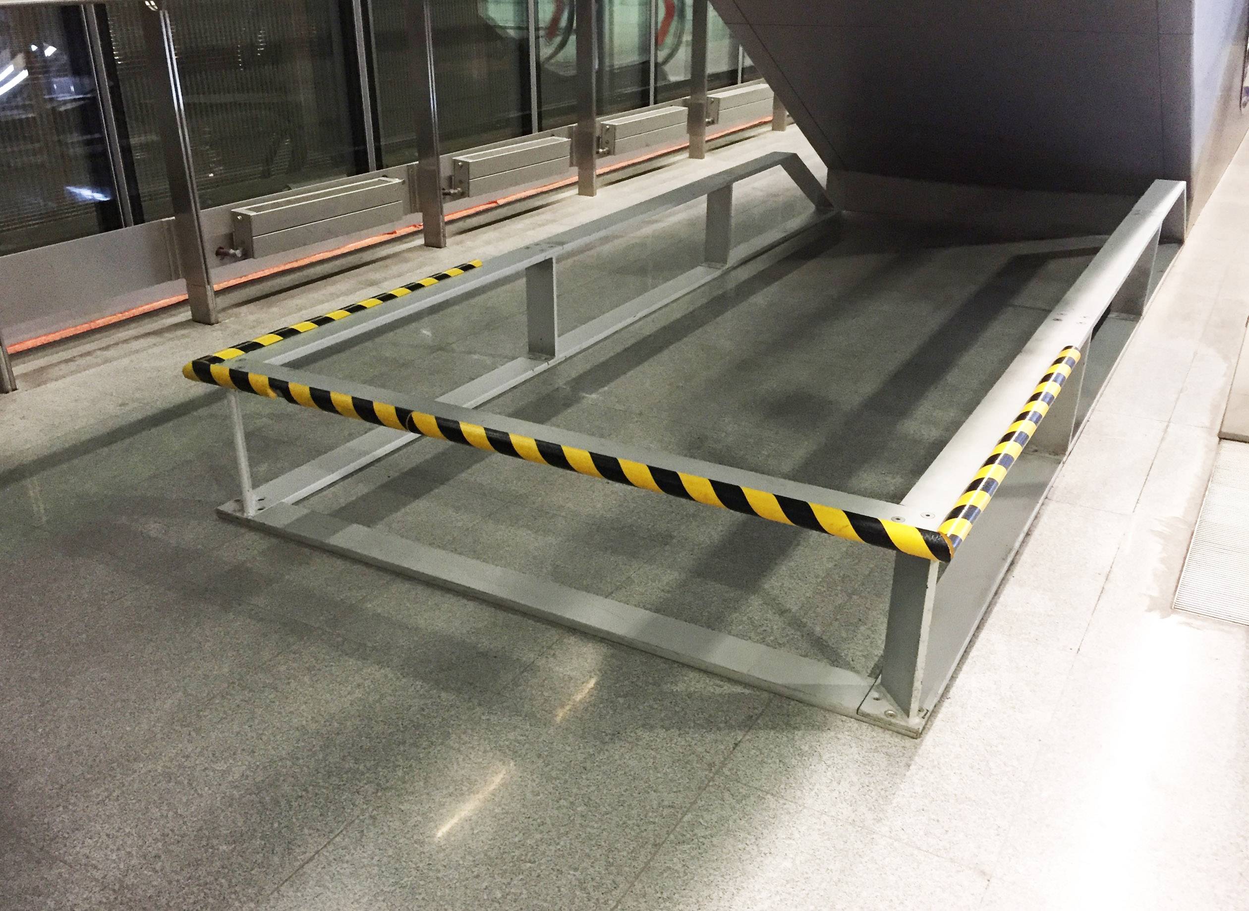 Metallgeländer mit gelb-schwarzem Warnstreifen umgibt eine kleine Treppenplattform in einer modernen, sauberen Umgebung.