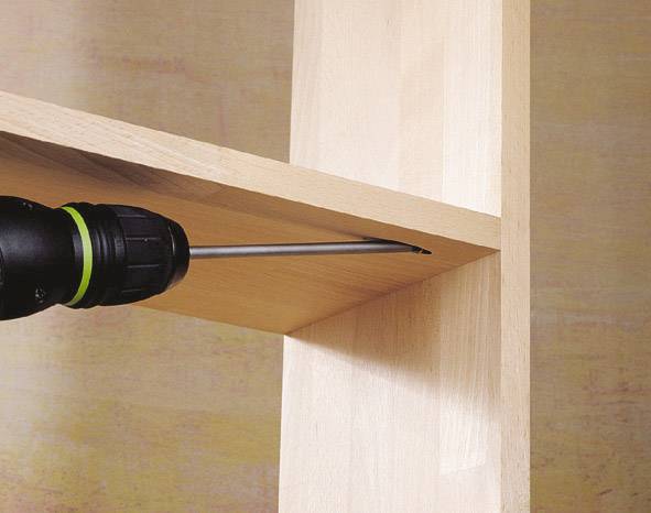 Eine Bohrmaschine schraubt ein Regalbrett an einen senkrechten Holzpfosten. Das Regal besteht aus hellem Holz.