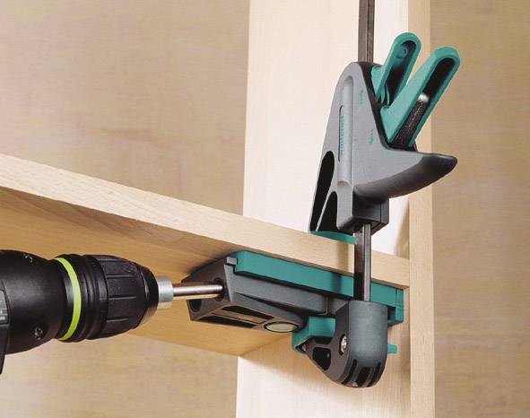 Eine Bohrmaschine mit Holzbohrer bohrt durch zwei Holzbretter, die mit einer grünen Spannzwinge fixiert sind.