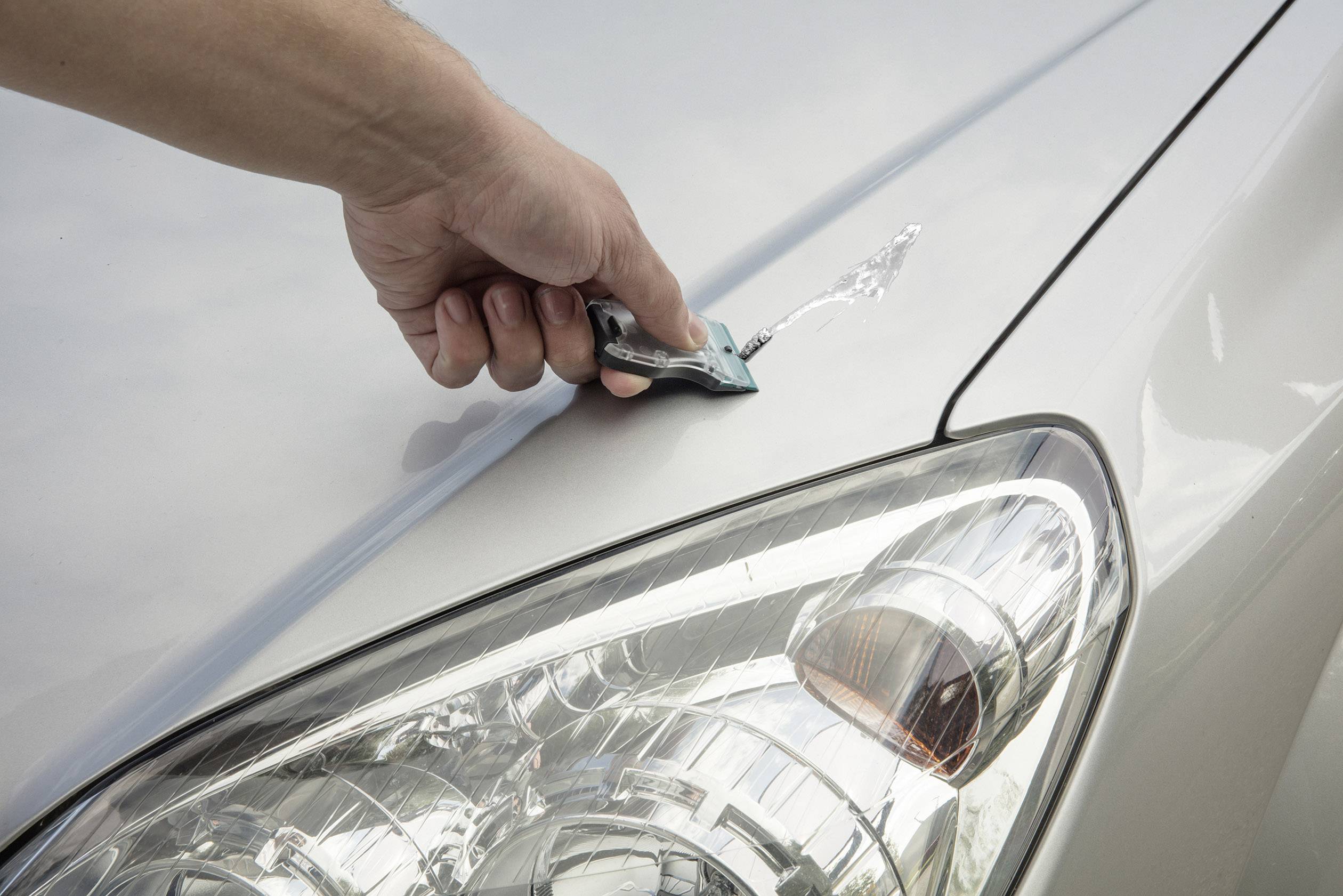 Hand mit Rasierklinge entfernt ein Aufkleber von der Motorhaube eines Autos. Vorderer Scheinwerfer ist im Bild zu sehen.