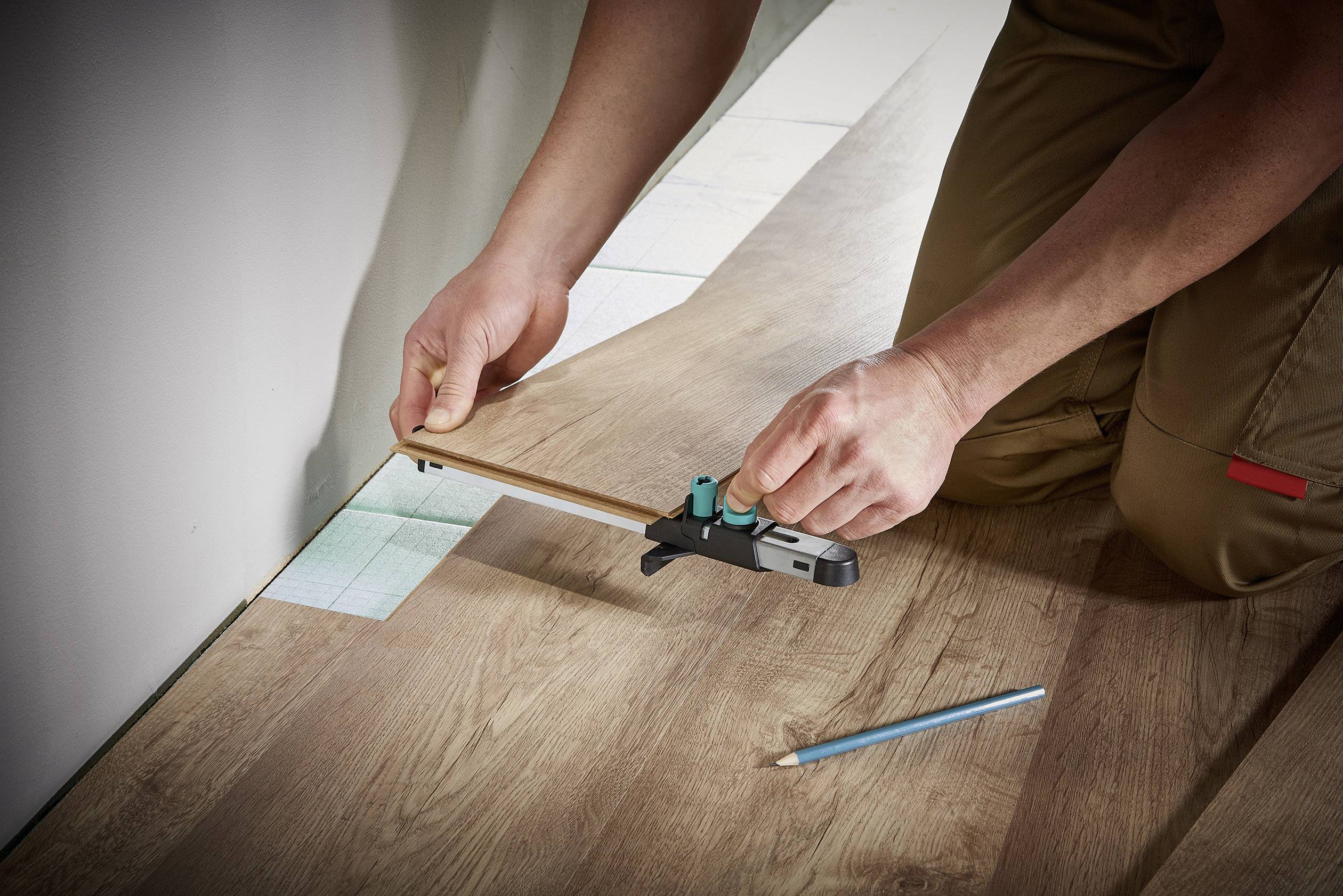 Person verlegt Laminatboden, verwendet ein Werkzeug zum genauen Messen und Schneiden. Ein Bleistift liegt daneben auf dem Boden.