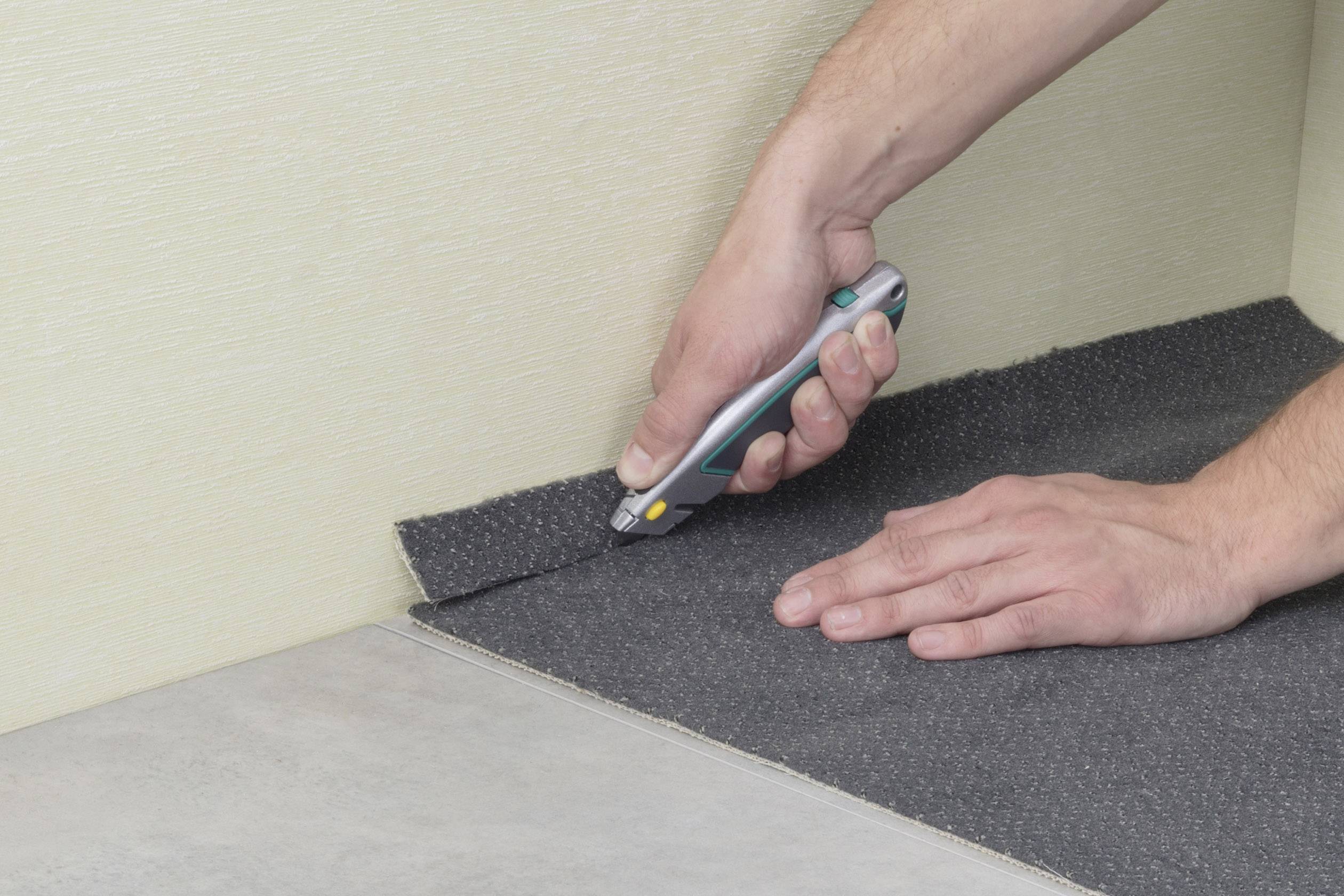 Eine Person schneidet mit einem Teppichmesser einen Teppich an der Wandkante passgenau zu. Hände fokussiert.