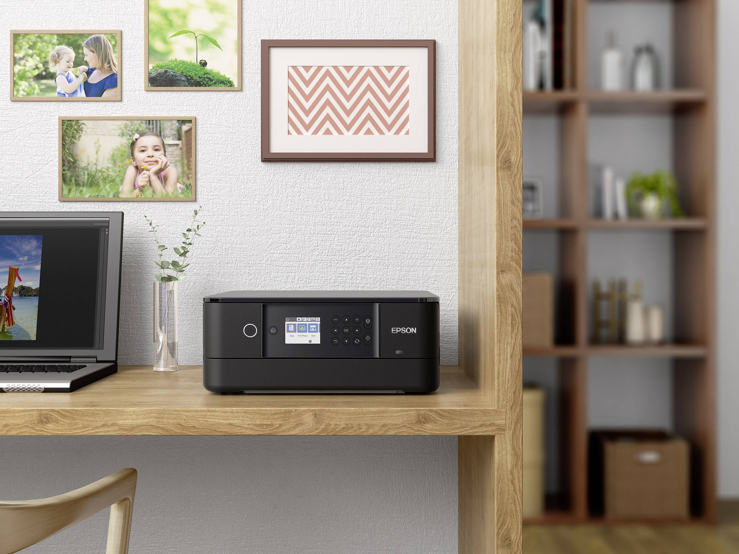Ein Drucker steht auf einem Schreibtisch neben einem Laptop, umgeben von gerahmten Fotos an der Wand und einem Regal im Hintergrund.