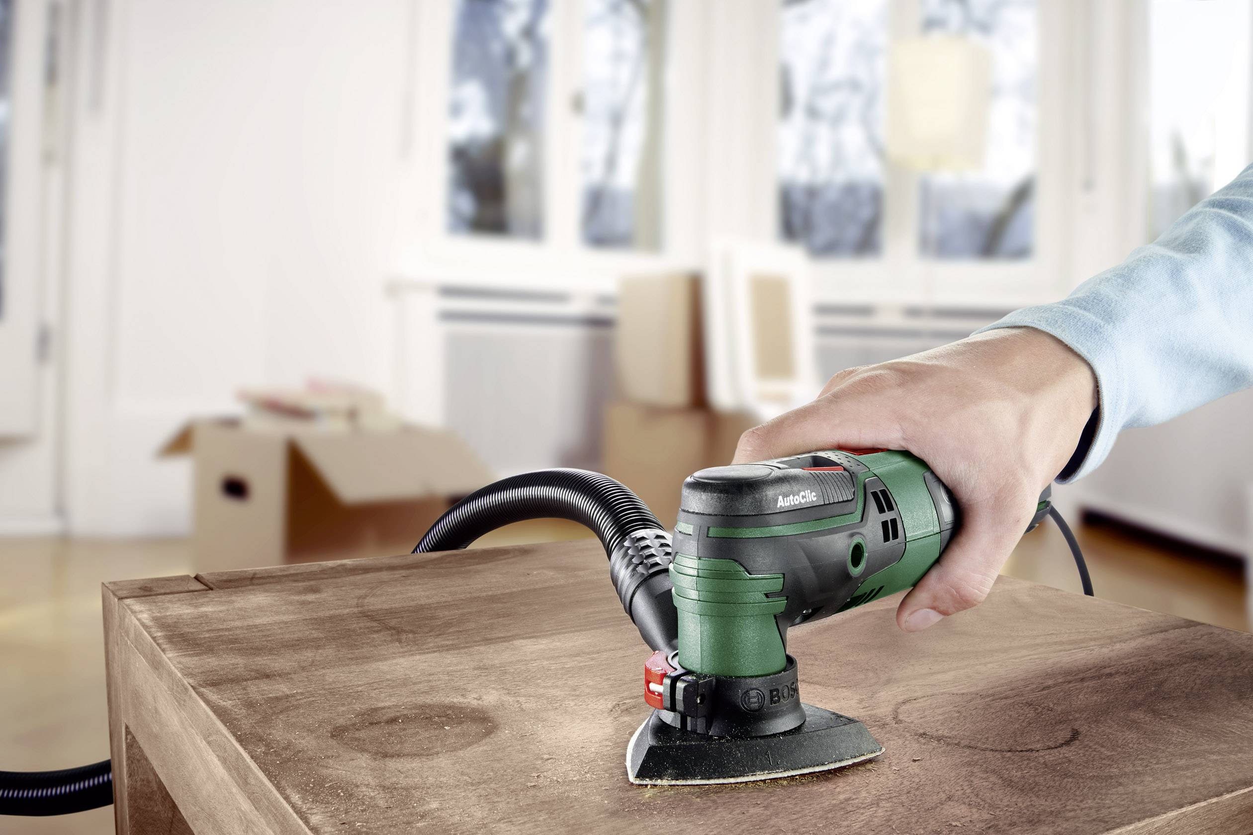 Eine Person schleift ein Holzbrett mit einem elektrischen Handwerkzeug in einem hellen Raum. Im Hintergrund sind Umzugskartons zu sehen.