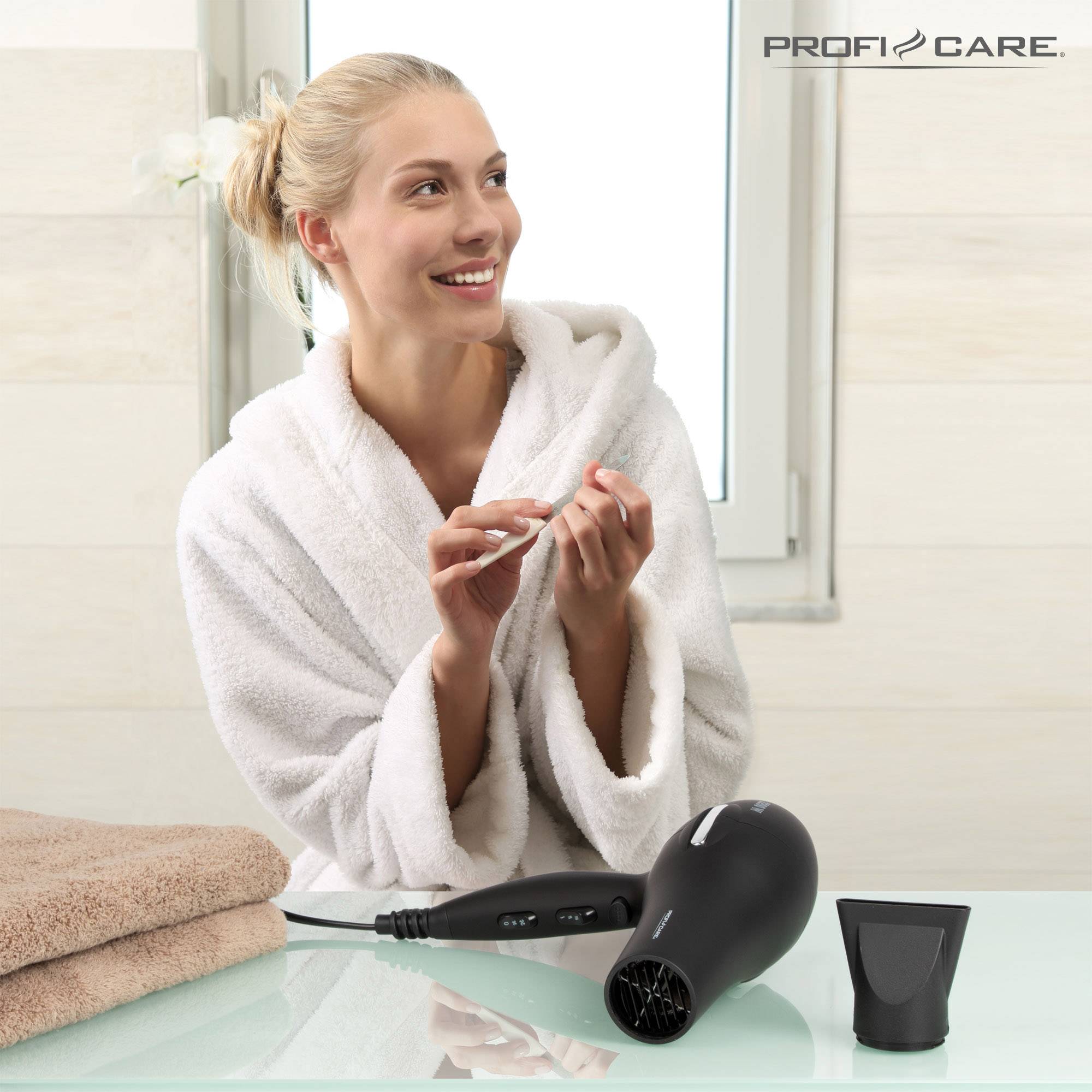 Eine Frau im Bademantel hält eine Gesichtscreme in einem Badezimmer. Auf dem Tisch liegt ein Föhn neben Handtüchern.