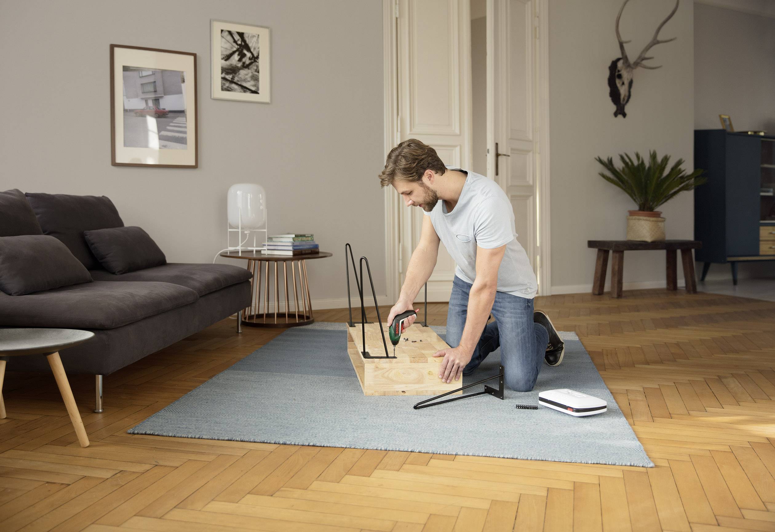Ein Mann montiert ein Möbelstück auf einem Teppich in einem Wohnzimmer. Im Hintergrund sind ein Sofa, Bilder und eine Pflanze sichtbar.
