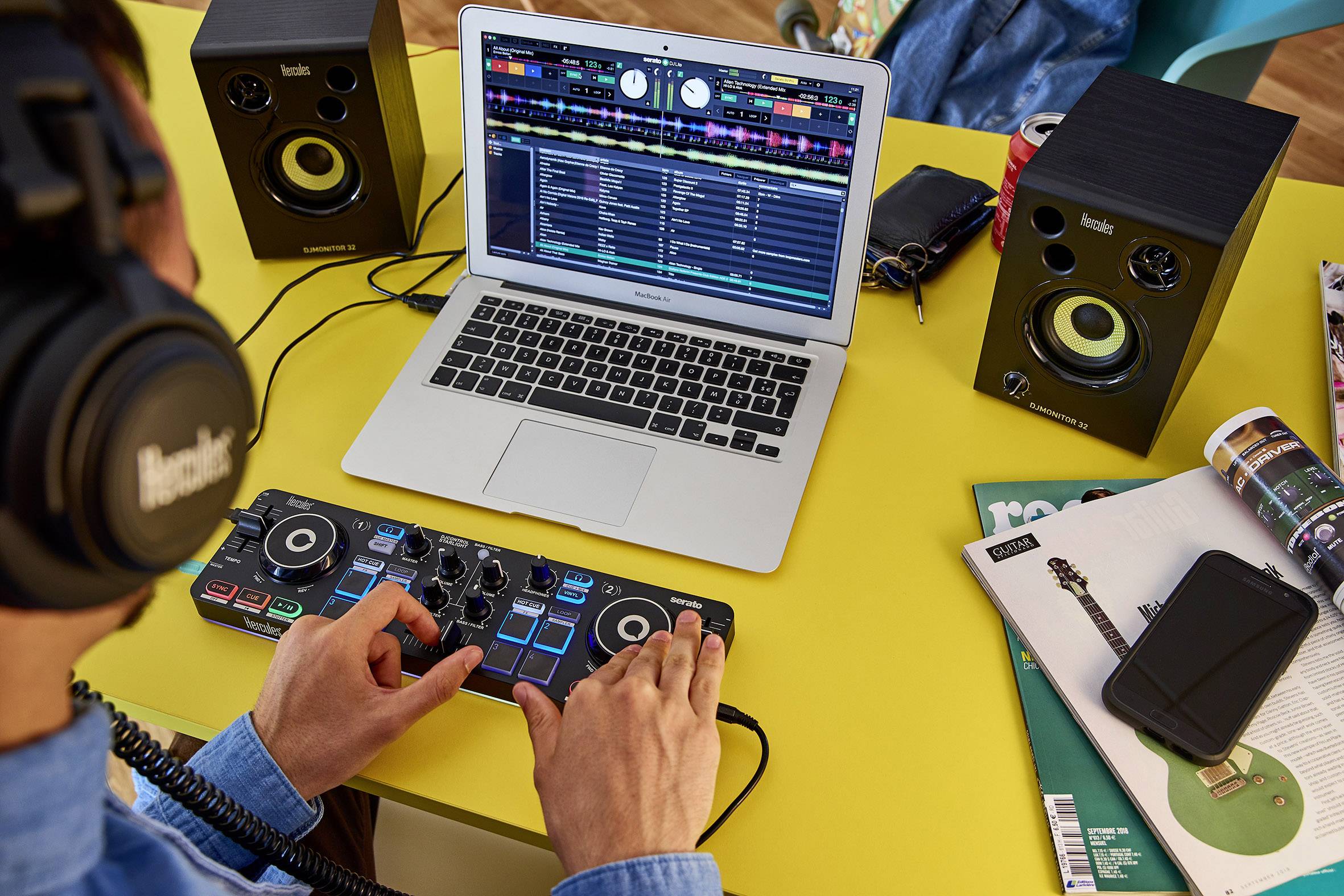 Ein Mann mit Kopfhörern sitzt an einem gelben Tisch. Vor ihm stehen ein Laptop, flankiert von zwei Lautsprechern, und ein kompakter DJ-Controller, den er gerade bedient. Darüber hinaus befinden sich eine Schlüsseltasche, eine Getränkedose, mehrere Zeitschriften und ein Smartphone auf dem Tisch.