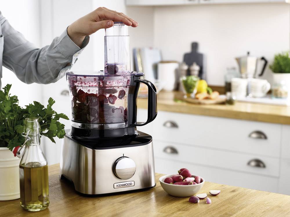 Eine Person benutzt eine Küchenmaschine, um rote Bete zuzubereiten. Die Maschine steht in einer modernen Küche mit Gemüsedekoration.