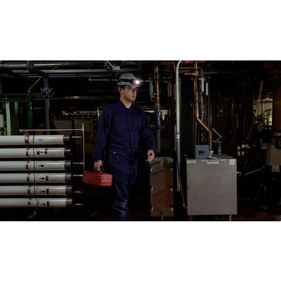 Ein Arbeiter in blauer Schutzkleidung und Helm trägt einen roten Werkzeugkasten in einem industriellen Raum mit Rohren und Maschinen.
