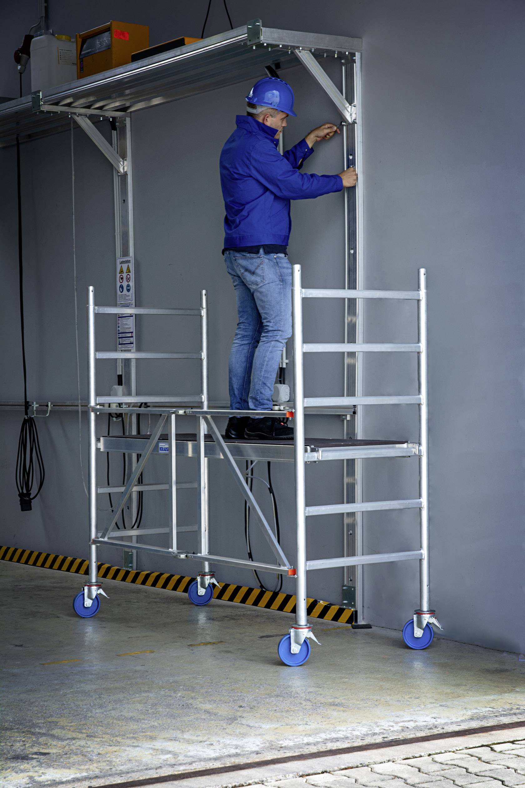 Ein Arbeiter steht auf einem mobilen Gerüst und arbeitet an einer Metallkonstruktion an der Wand. Sicherheitsausrüstung ist vorhanden.
