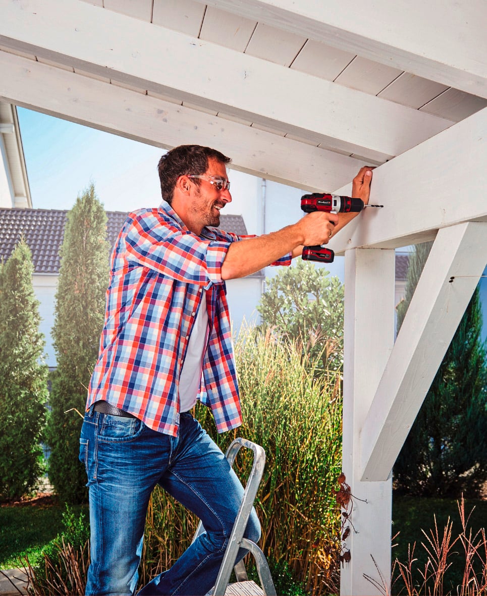 Ein Mann in kariertem Hemd und Jeans steht auf einer Leiter und bohrt mit einer elektrischen Bohrmaschine ein Loch in ein Holzgebälk.