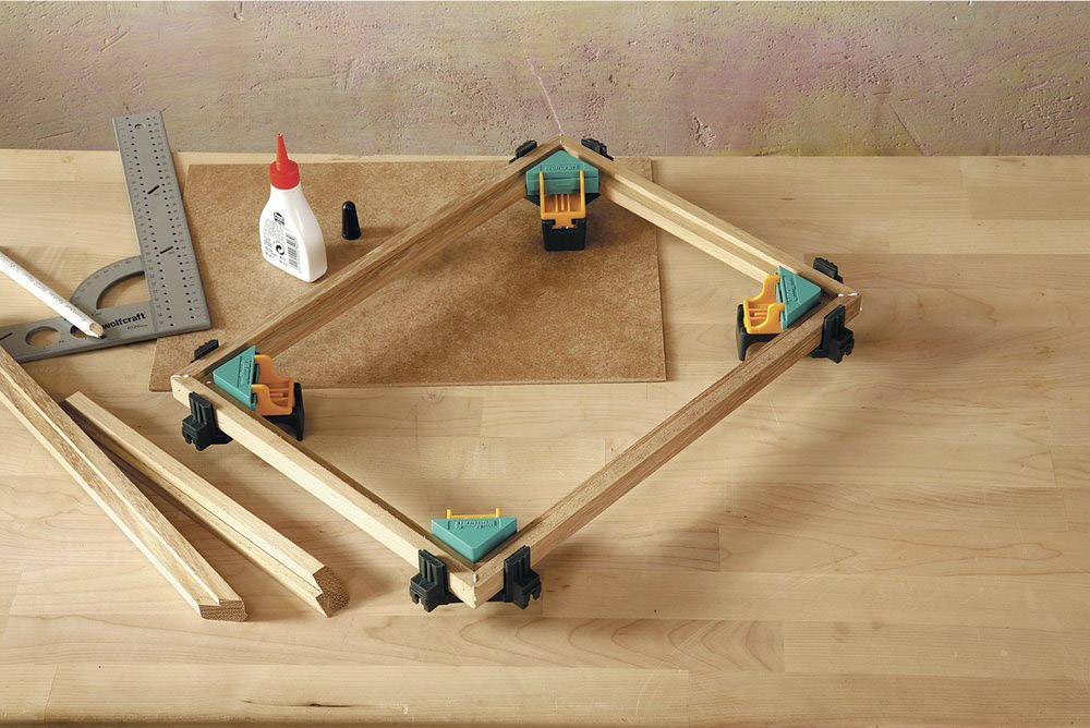Ein hölzerner Rahmen wird auf einer Werkbank gebaut, mit Klemmen gesichert. Auf dem Tisch liegen Winkelmesser und Leim.
