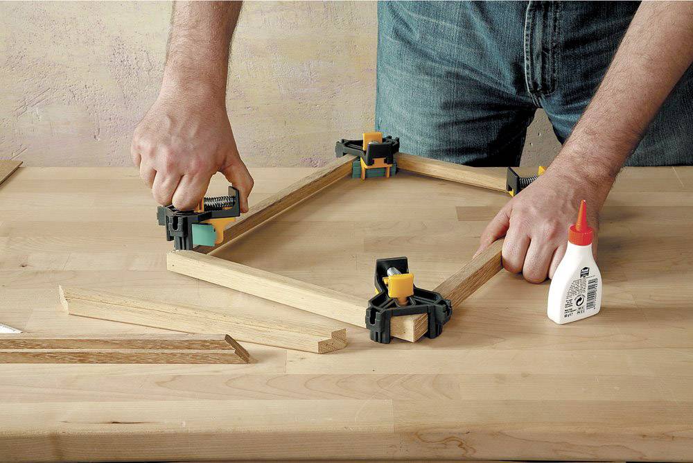 Eine Person baut einen Holzrahmen mit Eckzwingen und Holzleim auf einem Tisch zusammen. Hände positionieren und fixieren die Teile.
