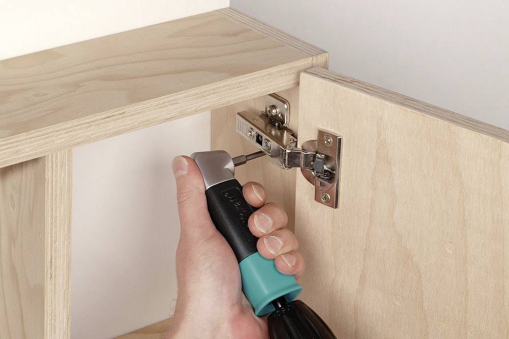 Eine Hand schraubt mit einem Schraubendreher ein Türscharnier an einen Holzschrank.