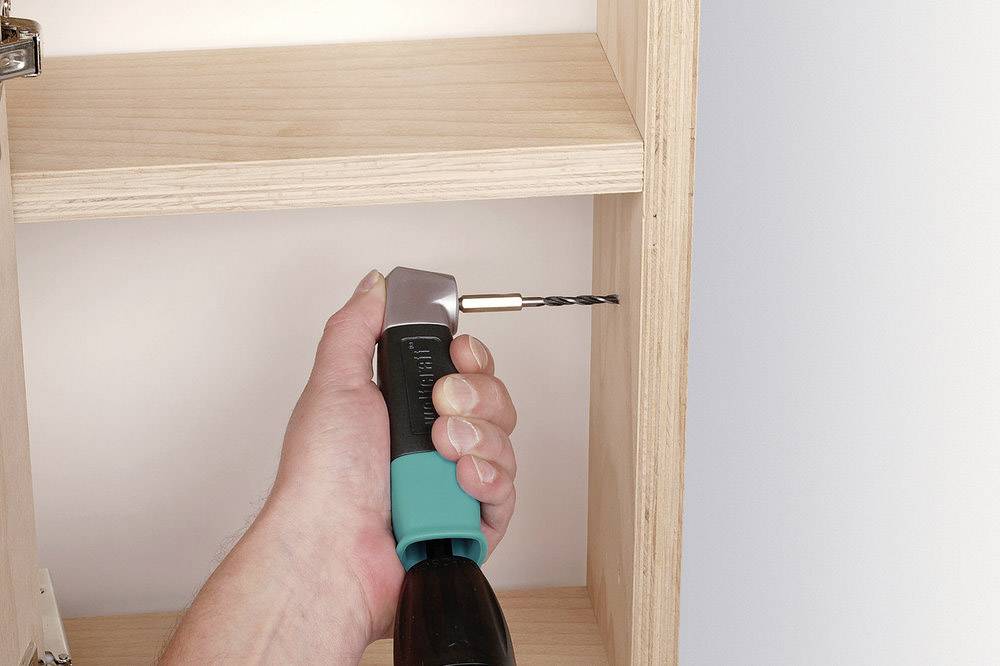 Eine Hand hält einen Bohrer mit Winkeladapter, der in ein Holzregal gebohrt wird. Der Adapter ermöglicht das Bohren in engen Räumen.