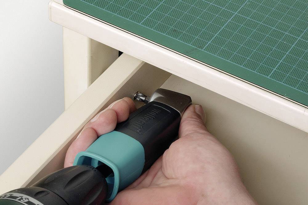 Eine Hand verwendet einen Akkuschrauber, um eine Schraube in einer Schublade anzuziehen. Oben links ist eine grüne Schneidematte.