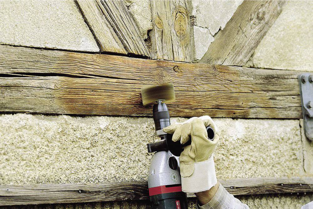 Handwerker schleift mit einer elektrischen Bürste Holz an einer Wand ab. Schutzbehandschuhe werden getragen.