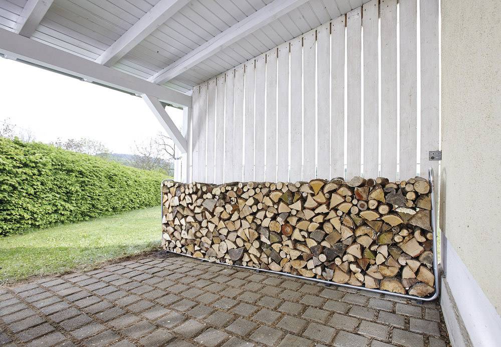 Ein Stapel Holzscheite liegt ordentlich gestapelt unter einem überdachten Carport auf einer gepflasterten Fläche neben einer Hecke.