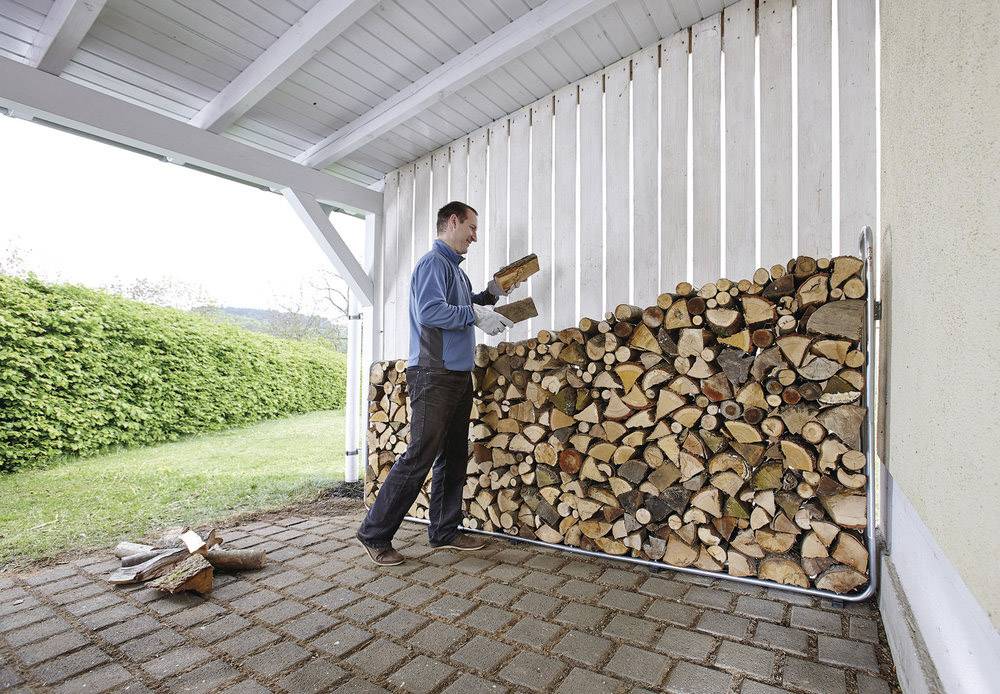 Ein Mann stapelt Brennholz unter einem überdachten Bereich. Links ist eine grüne Hecke zu sehen. Das Holz ist in einem Metallrahmen gestapelt.