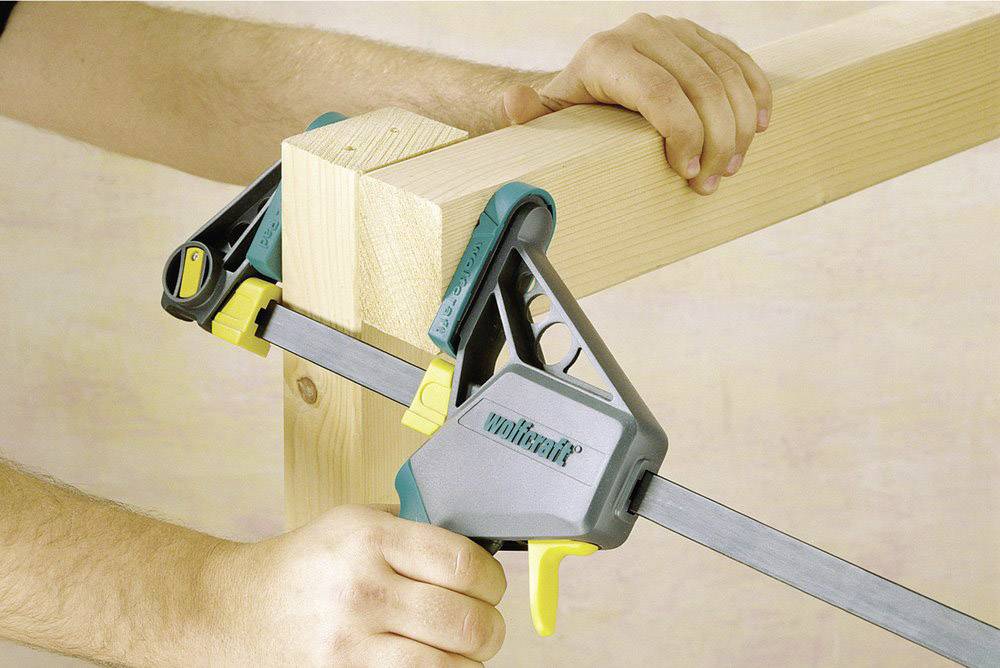 Eine Person verwendet eine Spannzwinge, um zwei Holzbalken im rechten Winkel zu fixieren. Die Spannzwinge ist grün und gelb.