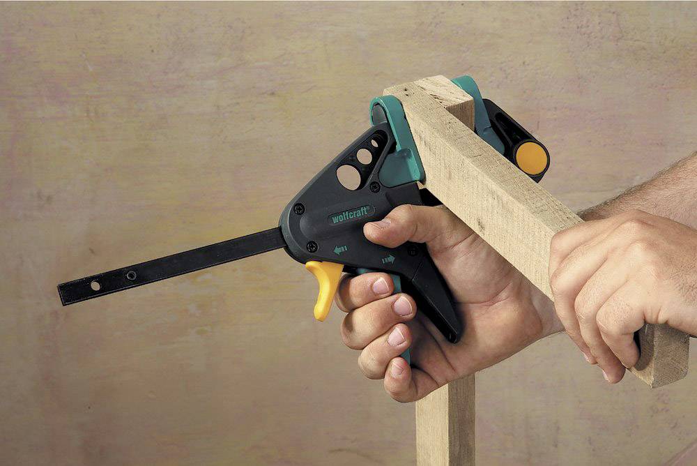 Eine Hand hält eine Einhandzwinge, die ein Holzstück fixiert. Die Zwinge ist schwarz mit einem gelben Griff und grünen Akzenten.