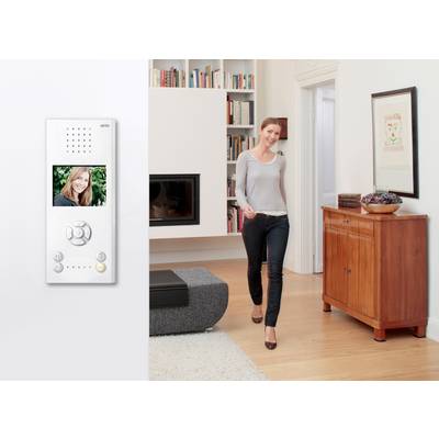 Eine Frau lächelt und geht durch ein modernes Wohnzimmer. An der Wand ein Intercom mit Display, das ein Bild derselben Frau zeigt.