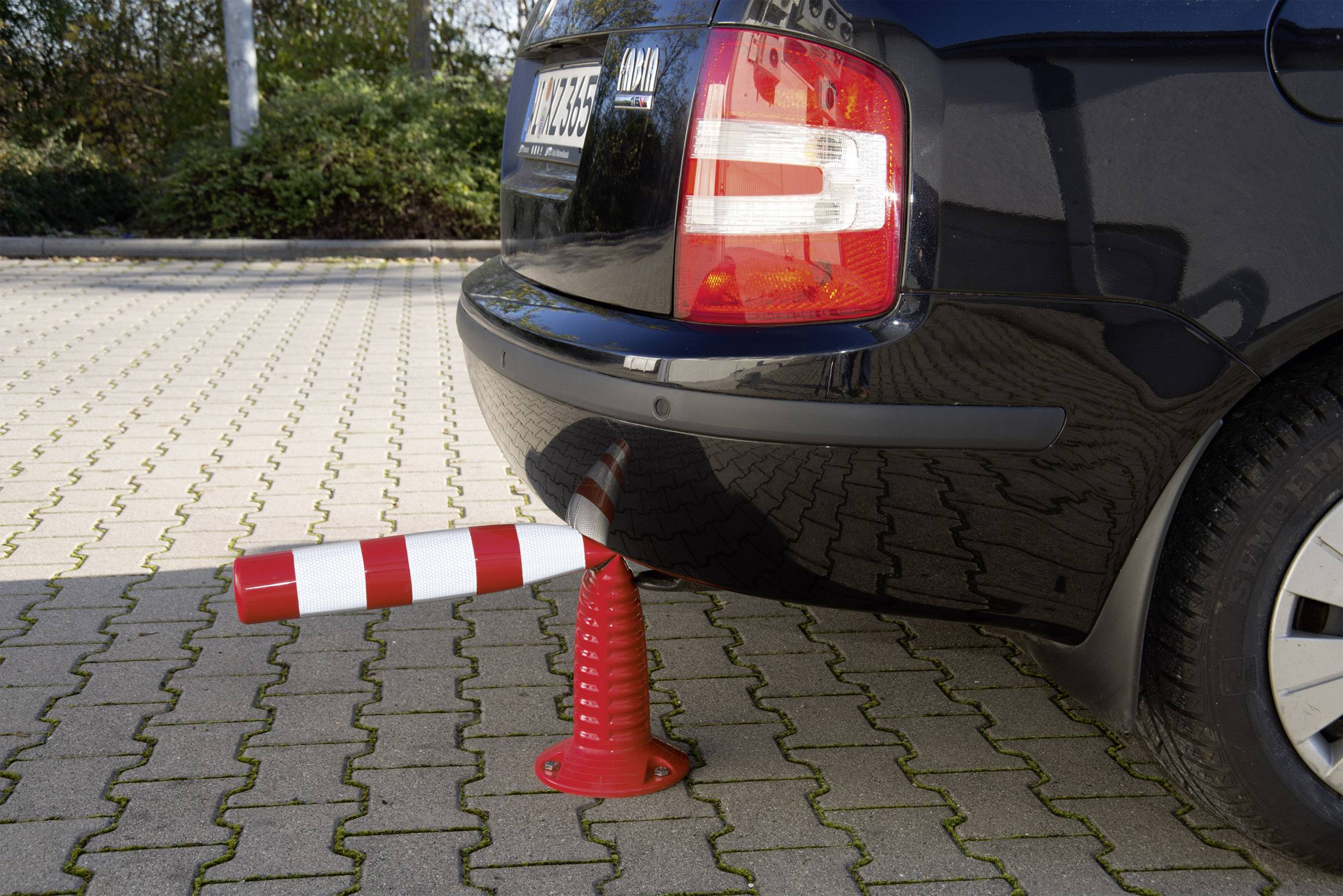 Ein Auto hat einen rot-weiß gestreiften Pfosten überfahren, der im hinteren Stoßfänger steckt. Der Pkw steht auf einem gemusterten Parkplatz.