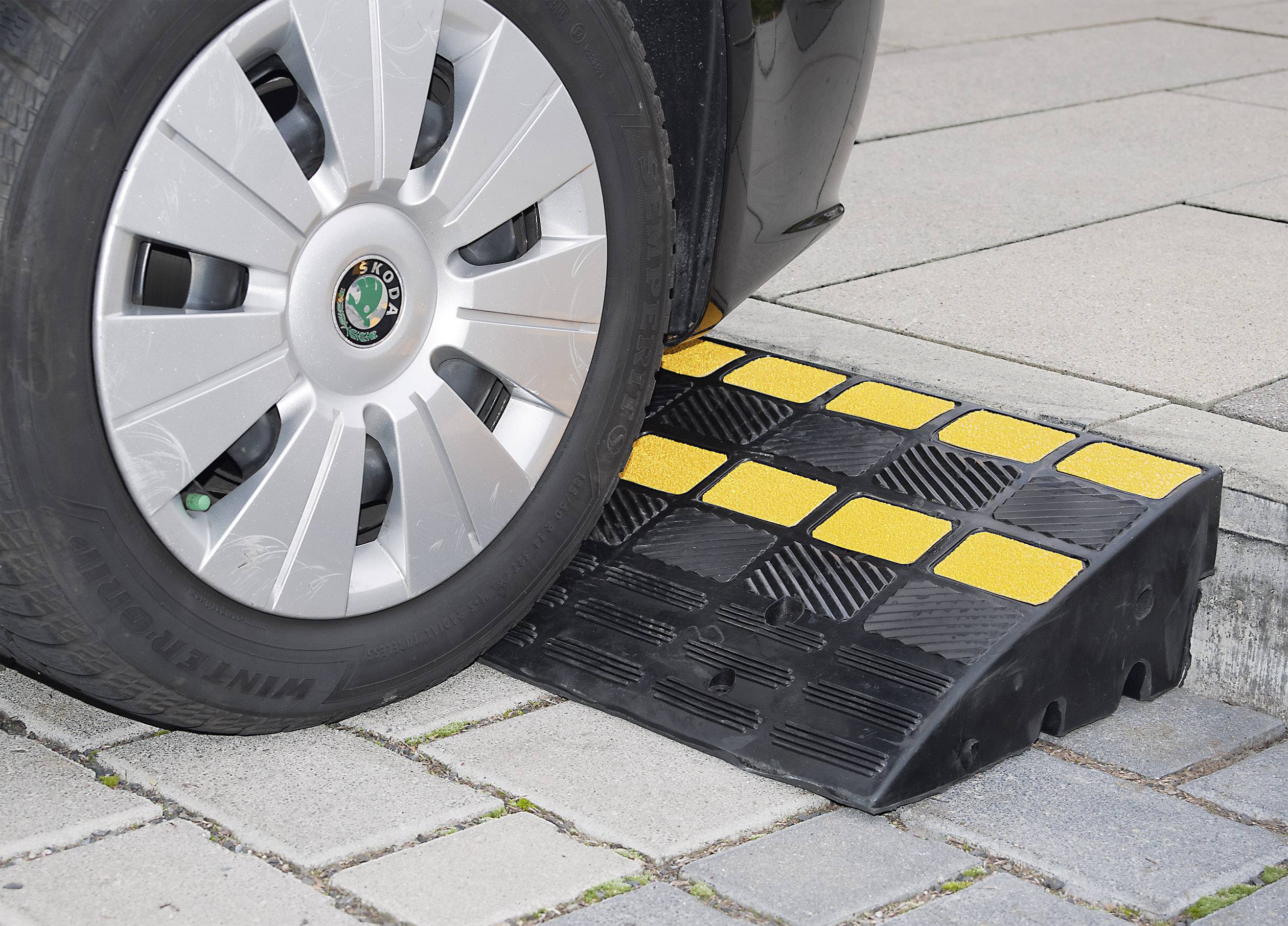 Ein Auto fährt über eine Bordsteinrampe aus schwarz-gelben Plastikmodulen auf gepflastertem Gehweg, um Höhenunterschied zu überwinden.