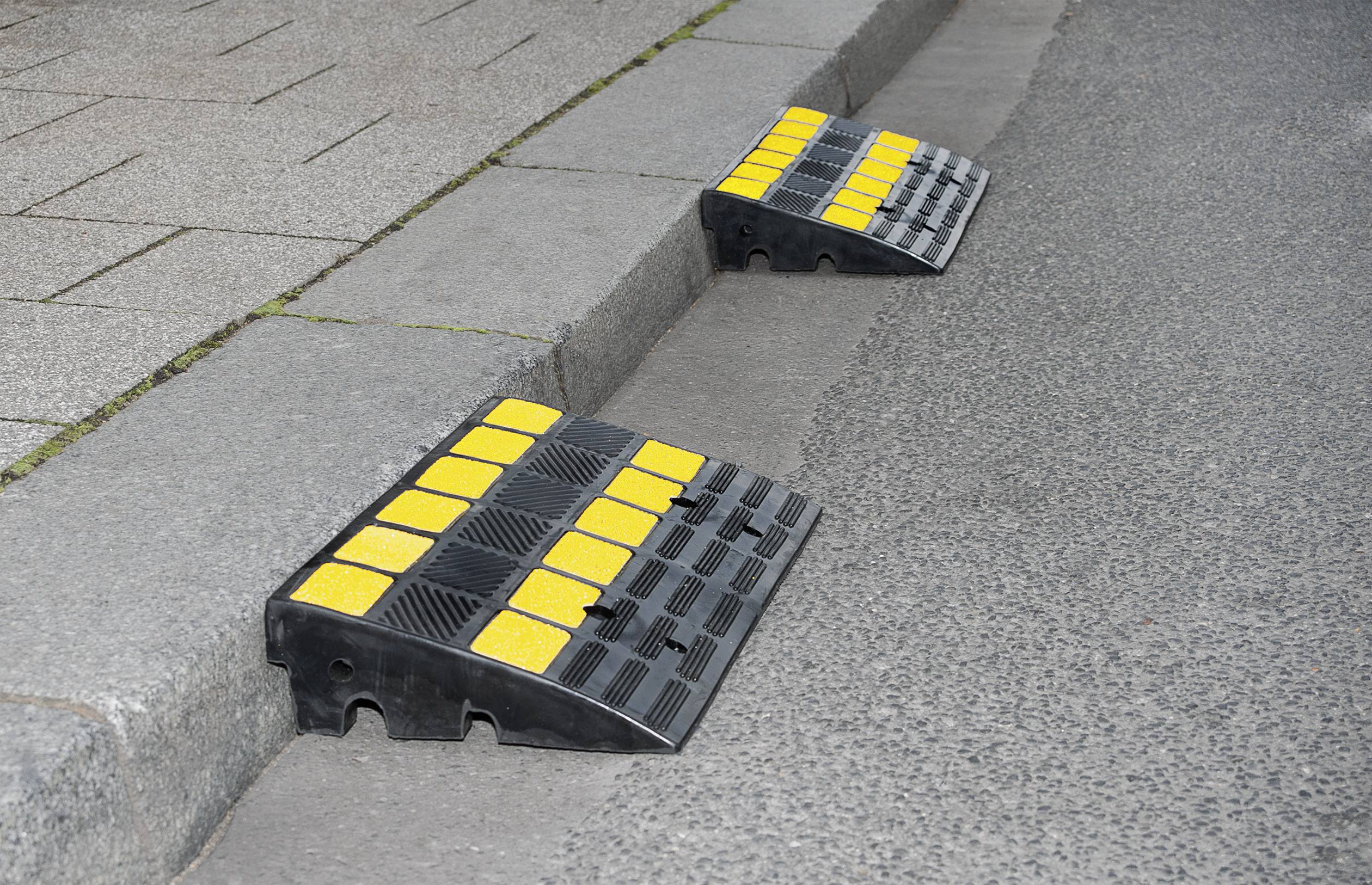 Schwarze und gelbe Kabelbrücke auf einer Bordsteinkante, die für Fahrzeuge passierbar ist und enge Fahrbahnen überwindet.