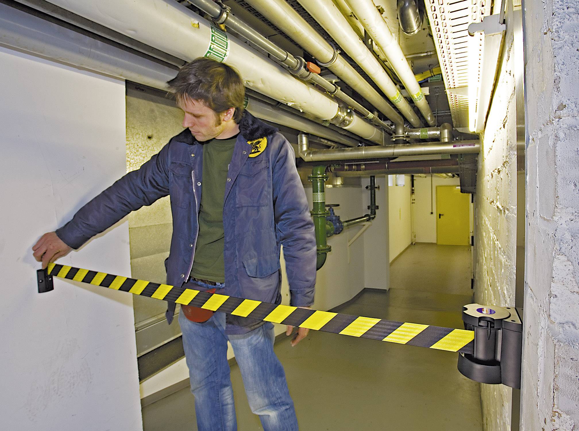 Ein Mann steht in einem technischen Kellerraum und spannt ein gelb-schwarzes Absperrband zwischen zwei Wänden.