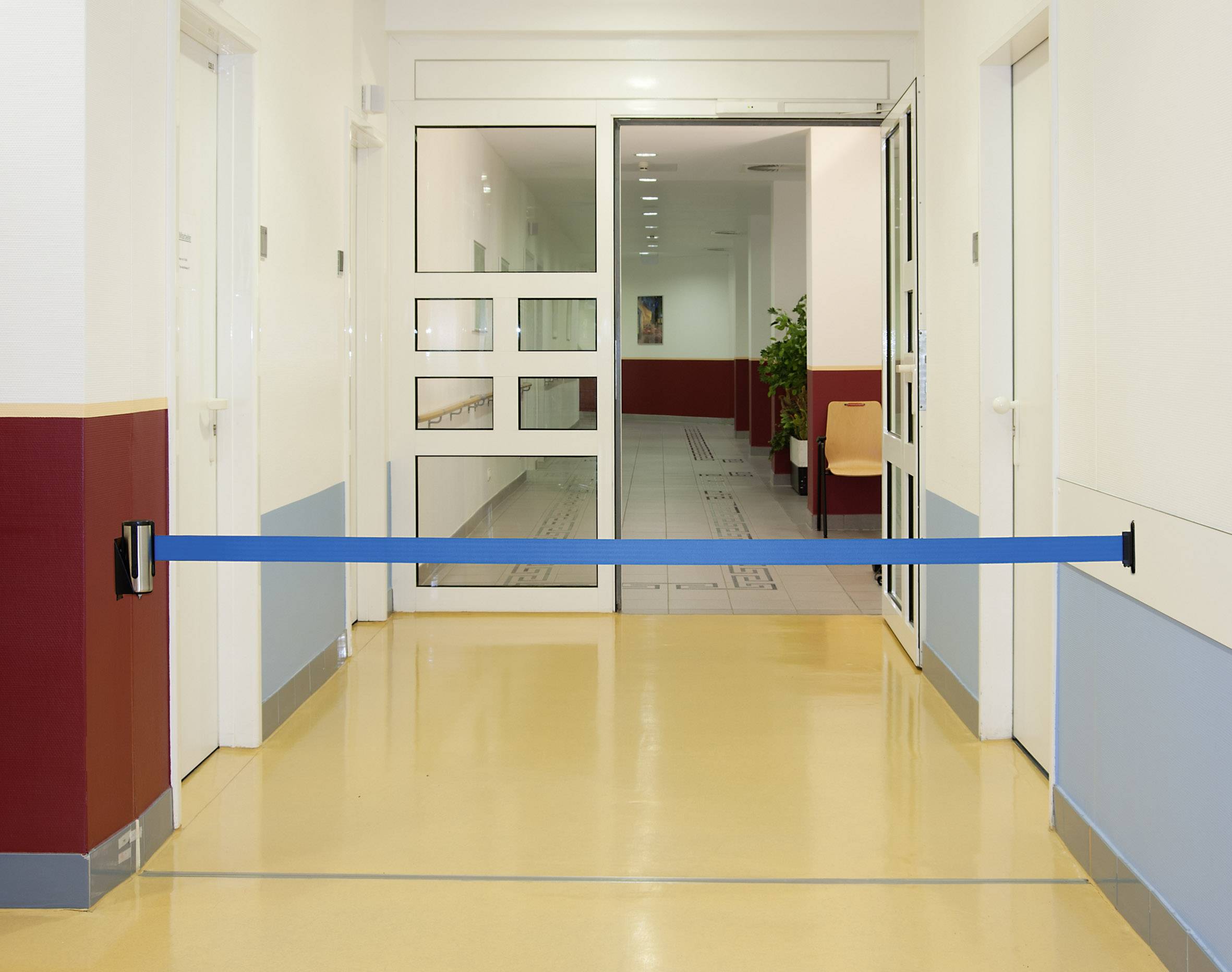 Ein Krankenhausflur mit versperrtem Zugang durch eine blaue Barriere, schließt eine Glastür ein und zeigt einen leeren Gang.