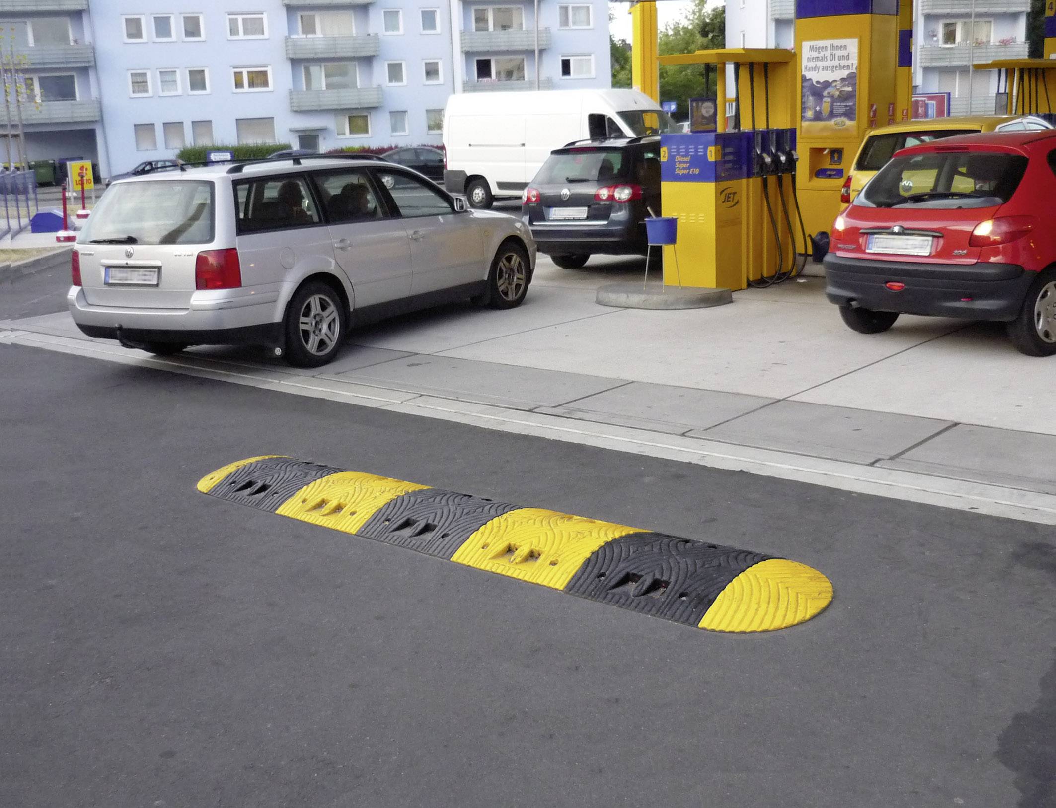 Autos an einer Tankstelle mit Bodenschwelle im Vordergrund zur Verkehrsberuhigung. Im Hintergrund sind Zapfsäulen sichtbar.