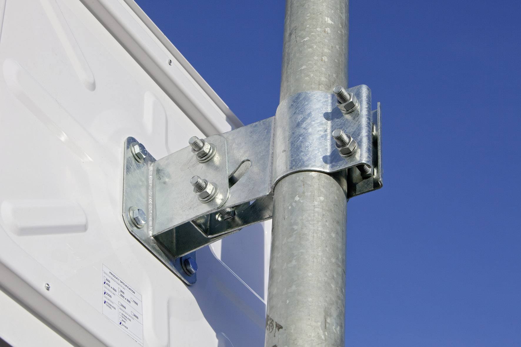 Ein Metallscharnier befestigt ein rechteckiges, weißes Schild an einem runden, grauen Metallpfosten vor klarem, blauem Himmel.