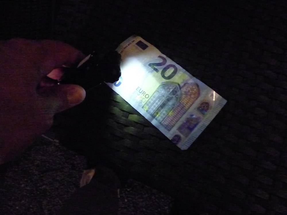 Eine Hand hält eine Taschenlampe über einen 20-Euro-Schein, um Sicherheitsmerkmale im Dunkeln zu prüfen.