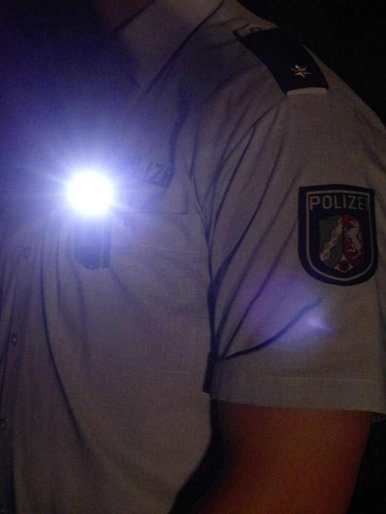 Person in Polizei-Uniform mit Schulterabzeichen. Eine Taschenlampe leuchtet an der Brust des Hemds. Beleuchtung und Uniform sind im Fokus.