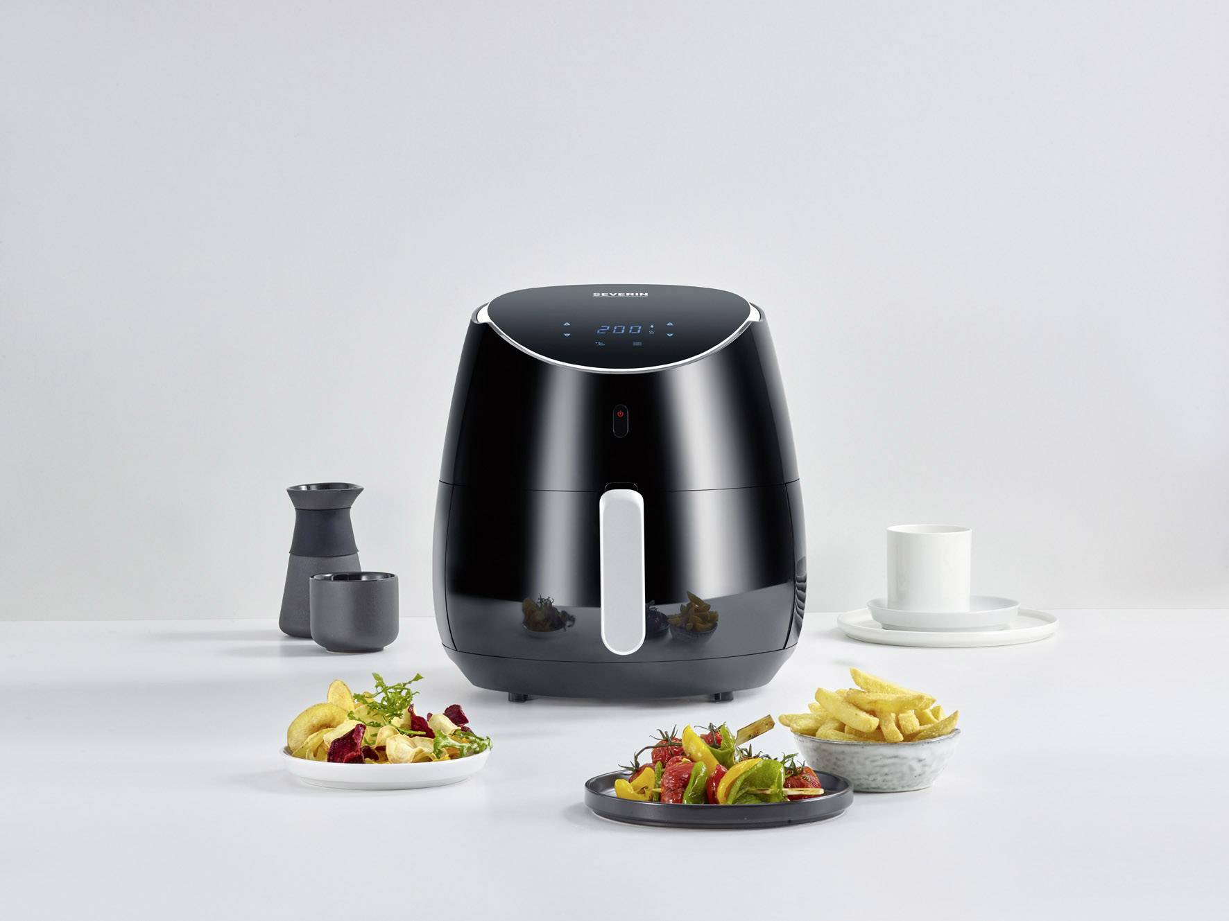 Schwarze Heißluftfritteuse auf einem Tisch mit verschiedenen Tellern mit Speisen und Getränken. Moderne Küchengeräteanordnung.