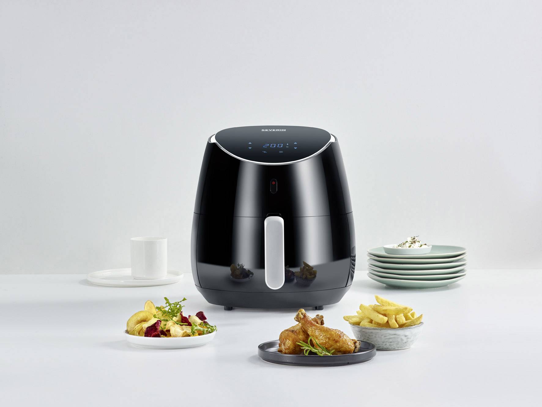 Ein schwarzer Airfryer mit digitalem Display steht auf einem Tisch, umgeben von Tellern mit frittierten Speisen und Geschirr.