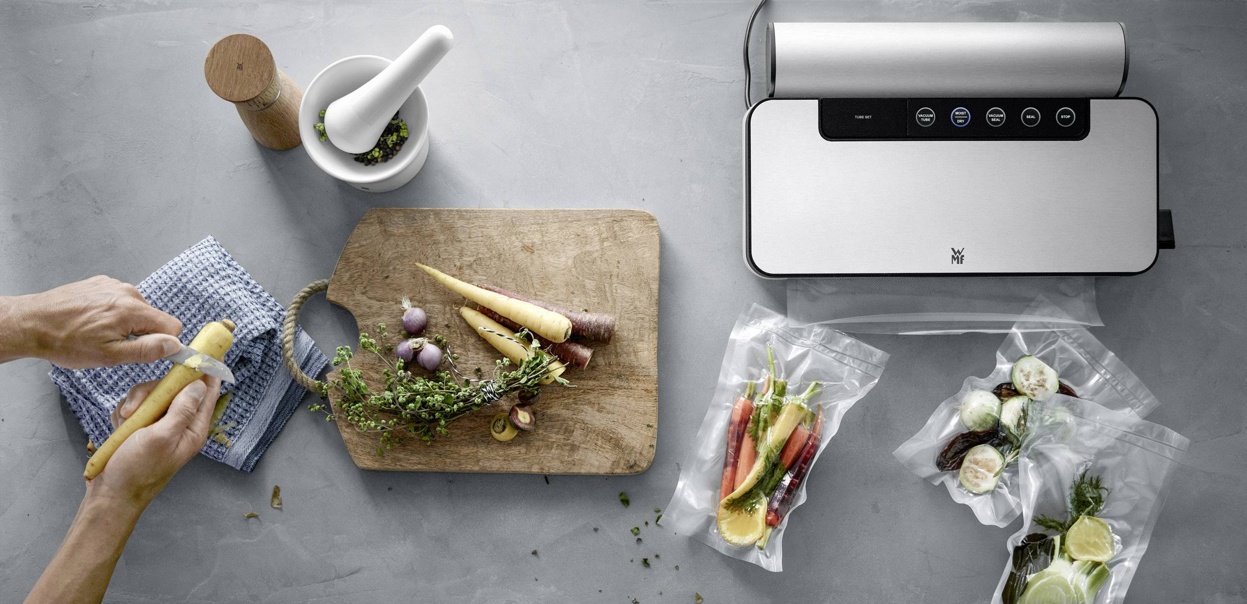Eine Person schält Karotten neben einem Schneidebrett mit Gemüse. Ein Vakuumiergerät versiegelt Lebensmittel in Plastikbeuteln.