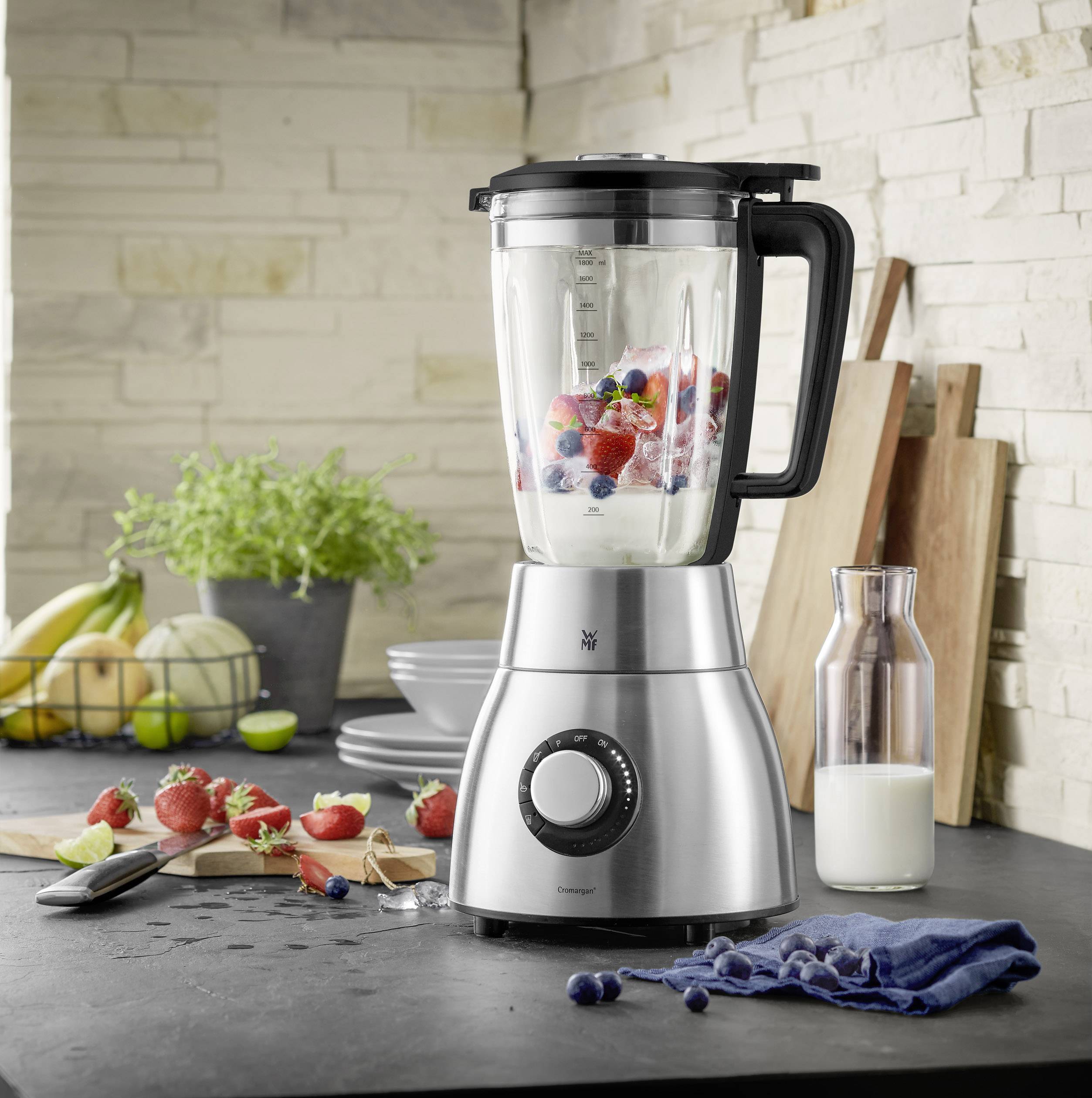 Ein silberner Mixer auf einer Küchenarbeitsplatte, gefüllt mit Beeren. Im Hintergrund sind Obst, Kräuter und Küchenutensilien zu sehen.