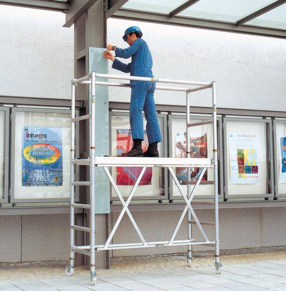 Ein Arbeiter in blauer Arbeitskleidung steht auf einem mobilen Gerüst und installiert oder repariert etwas an einer Wand.