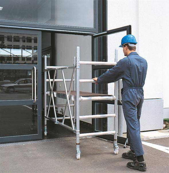 Ein Arbeiter in Schutzkleidung schiebt ein fahrbares Gerüst durch eine offene Tür eines Gebäudes. Der Eingangsbereich ist modern gestaltet.